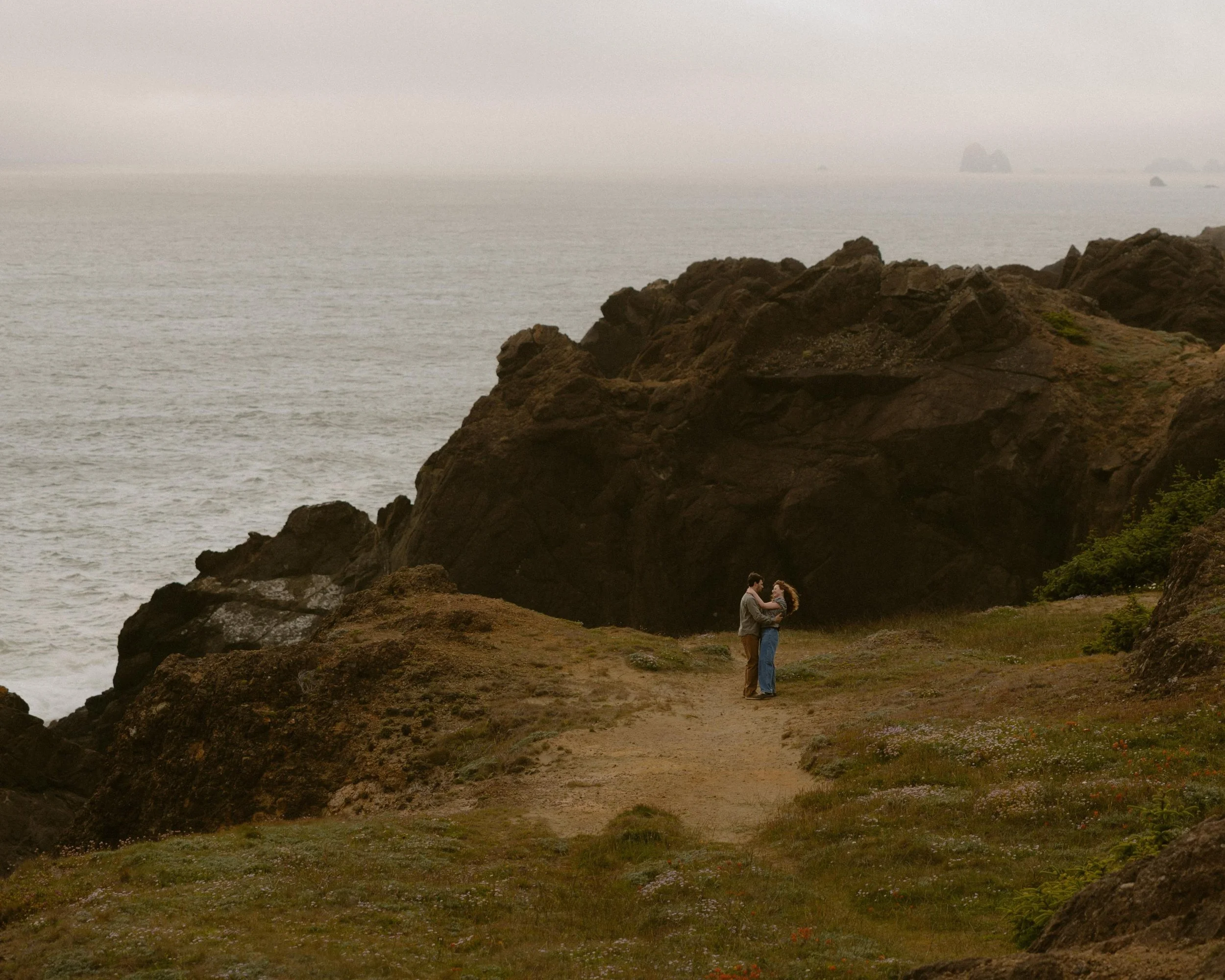 A couple stands on a rocky coastal area, holding each other, with the ocean and large rocks in the background on a foggy day.