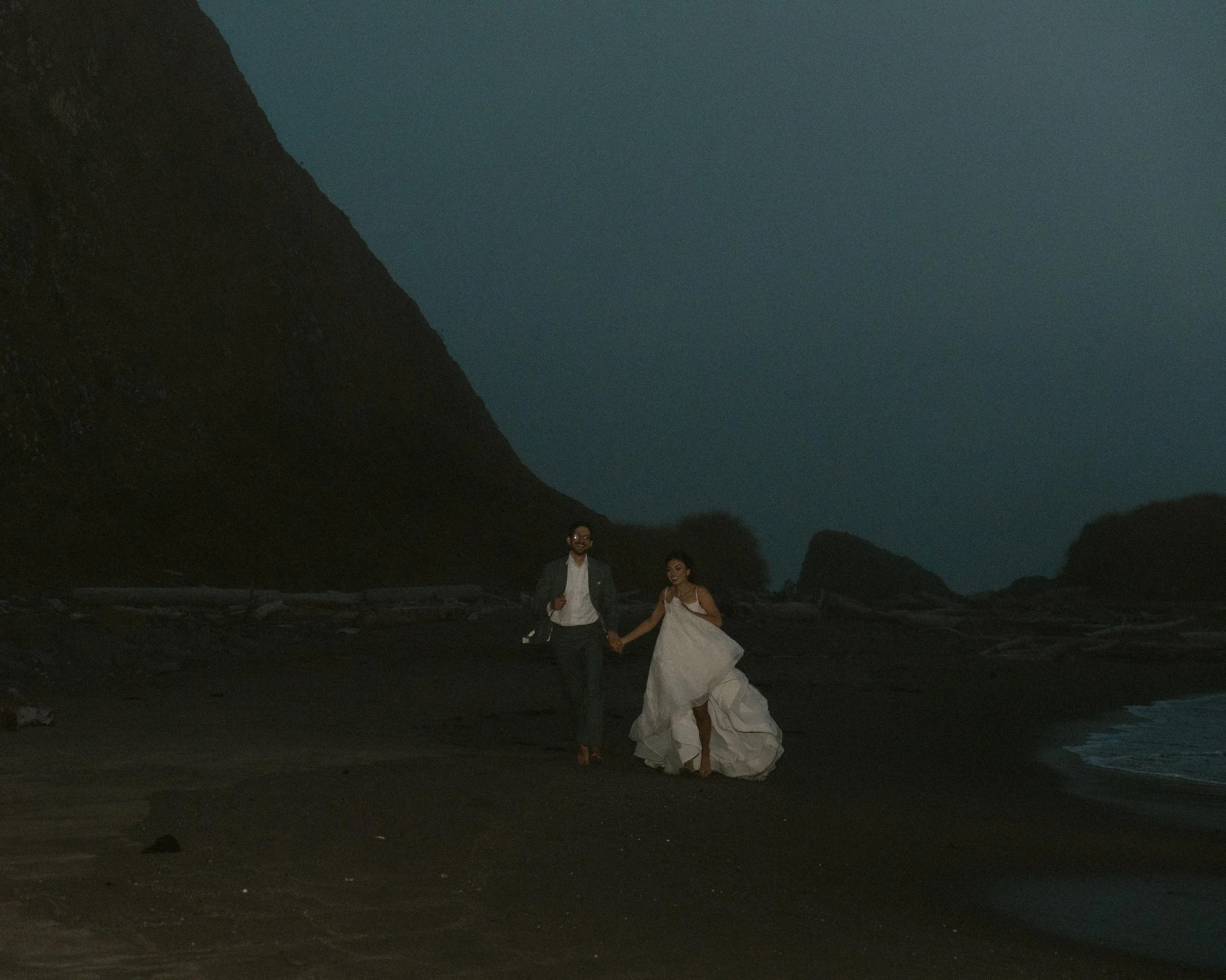 Nighttime scene at the beach with two people holding hands, one in a white dress and the other in dark clothing, near large rock formations and a mountain with dense greenery in the background.