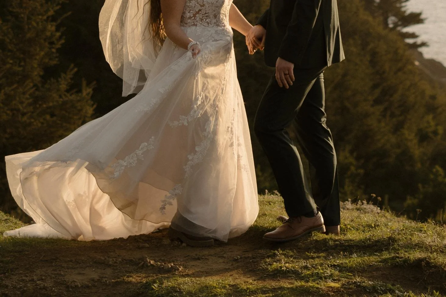 A bride and groom holding hands outdoors during sunset, standing on a grassy area with trees and a body of water in the background.