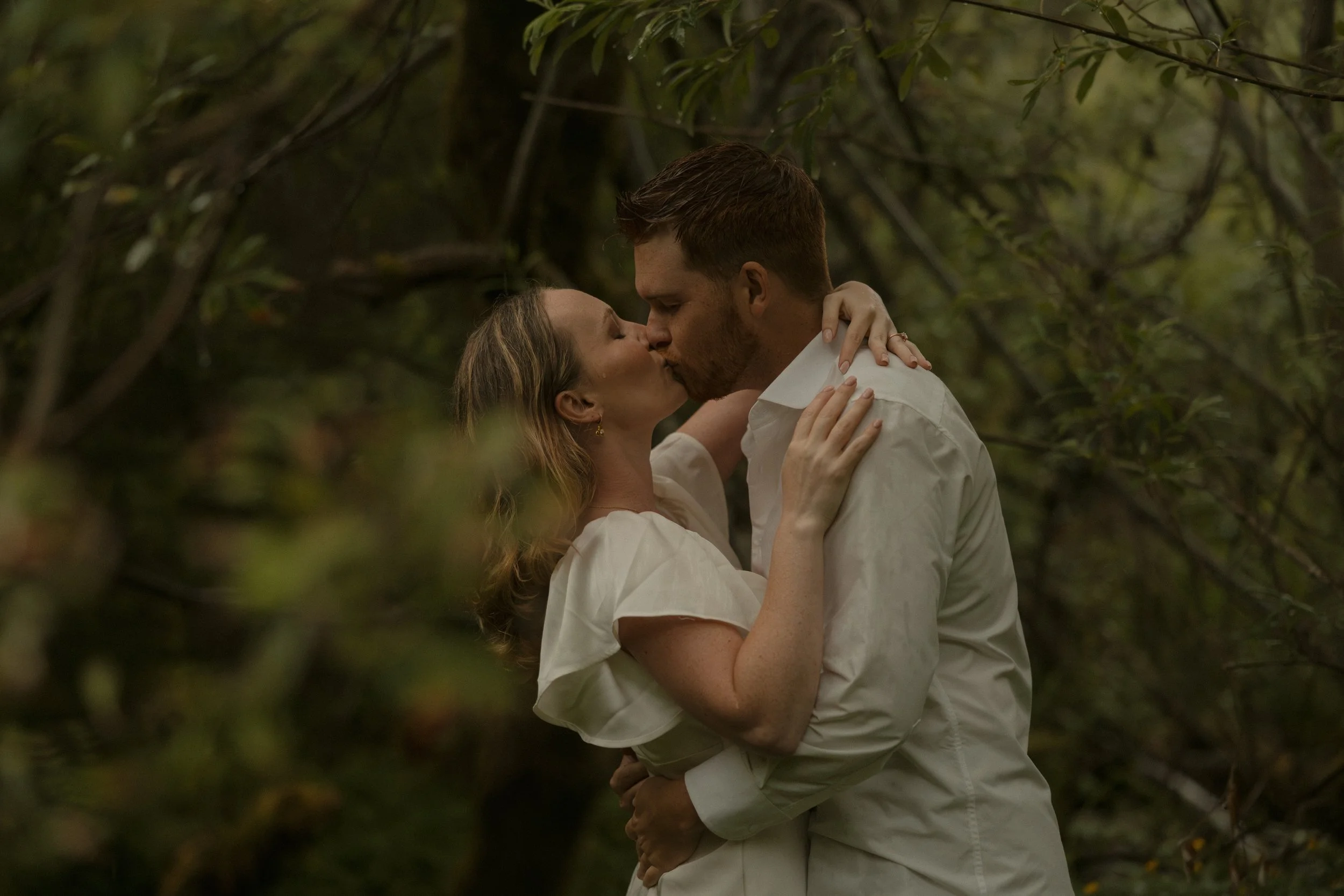 A couple sharing a kiss outdoors surrounded by trees and greenery.