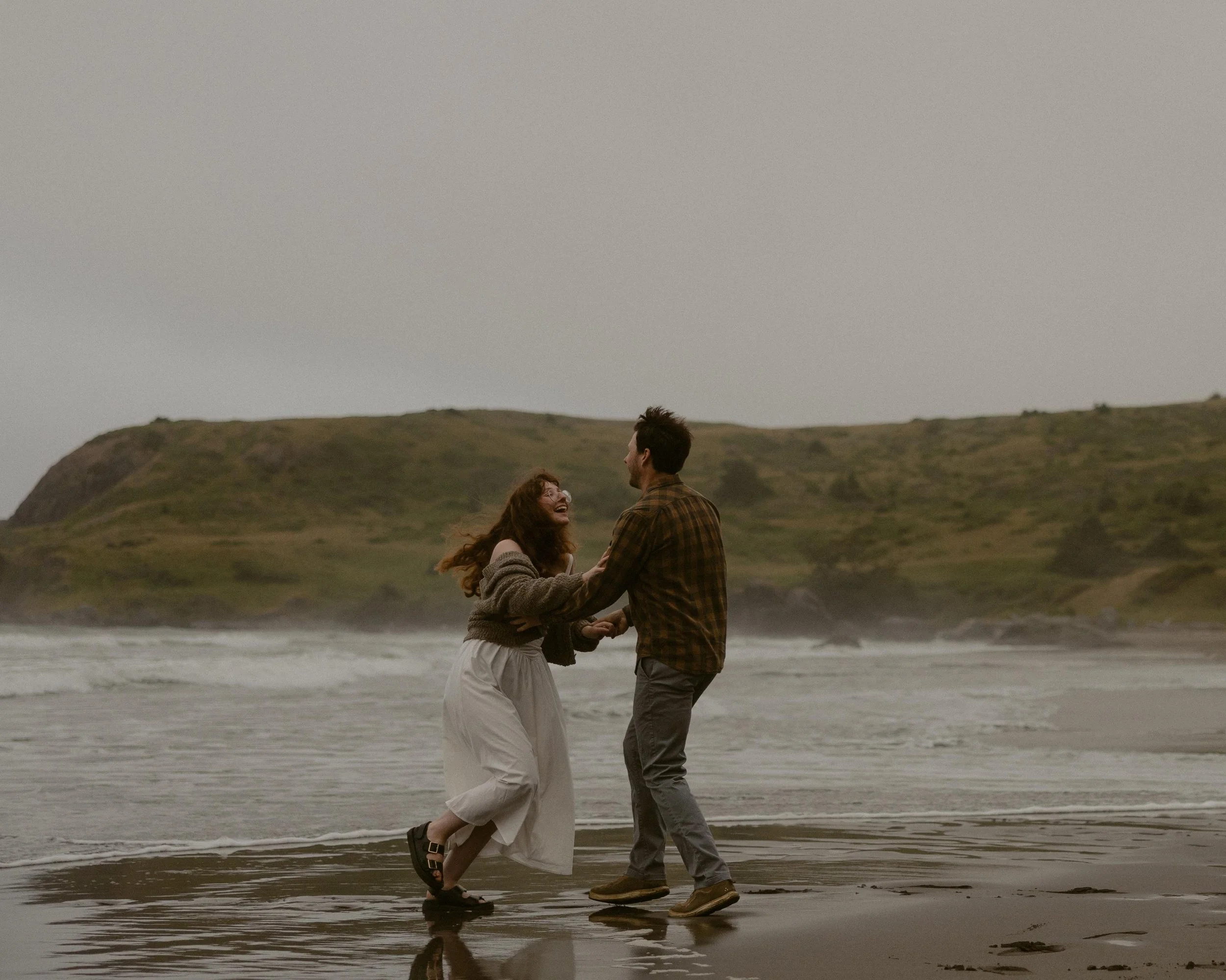 A couple is standing on the beach holding hands and looking at each other, with hills in the background and the ocean behind them.