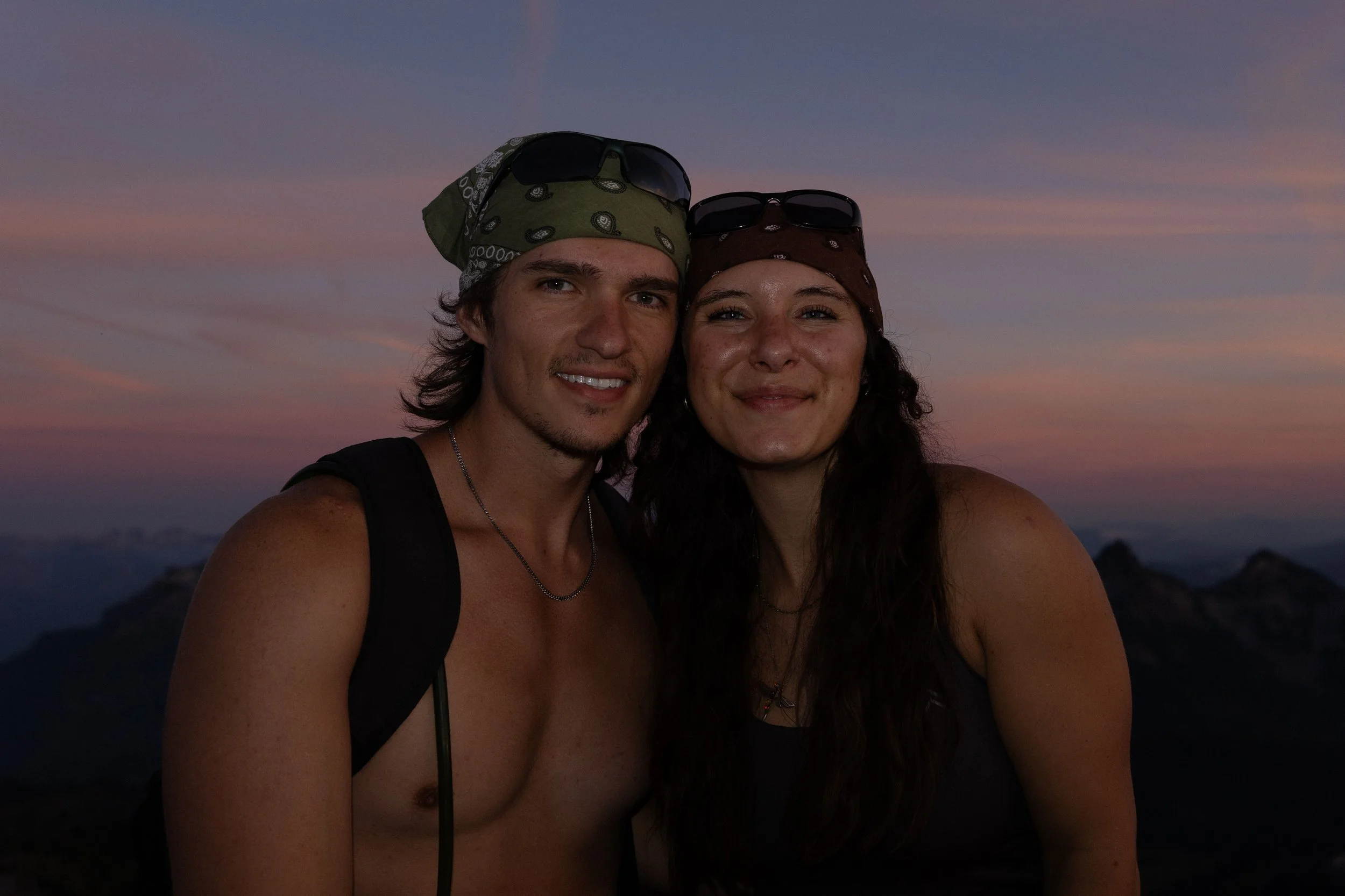 A young man and woman taking a selfie together outdoors at sunset, with mountain peaks visible in the background. They are wearing bandanas on their heads and sunglasses, and the woman is topless.