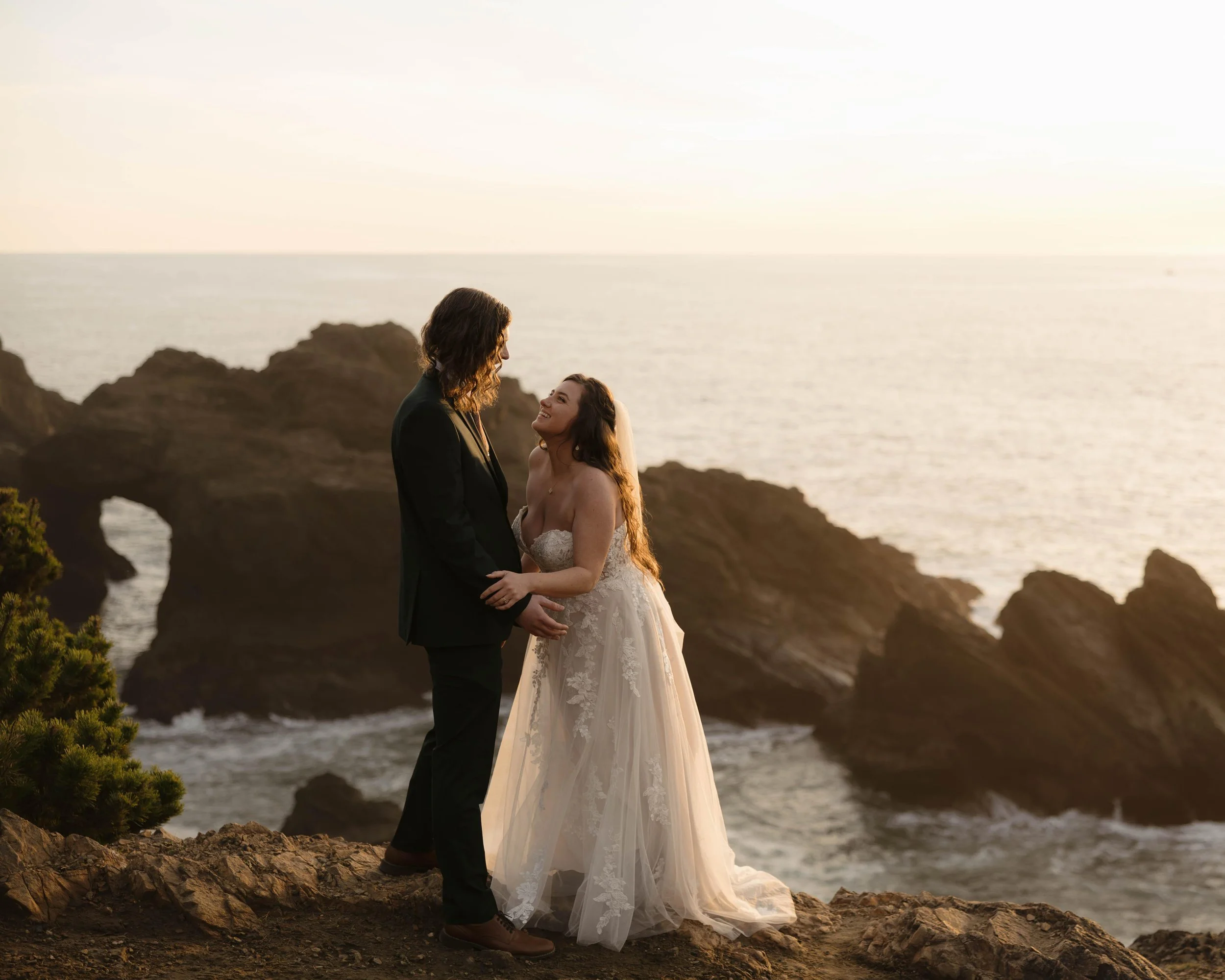A couple dressed in wedding attire standing on rocks by the ocean at sunset, gazing at each other with rocky cliffs and the sea in the background.