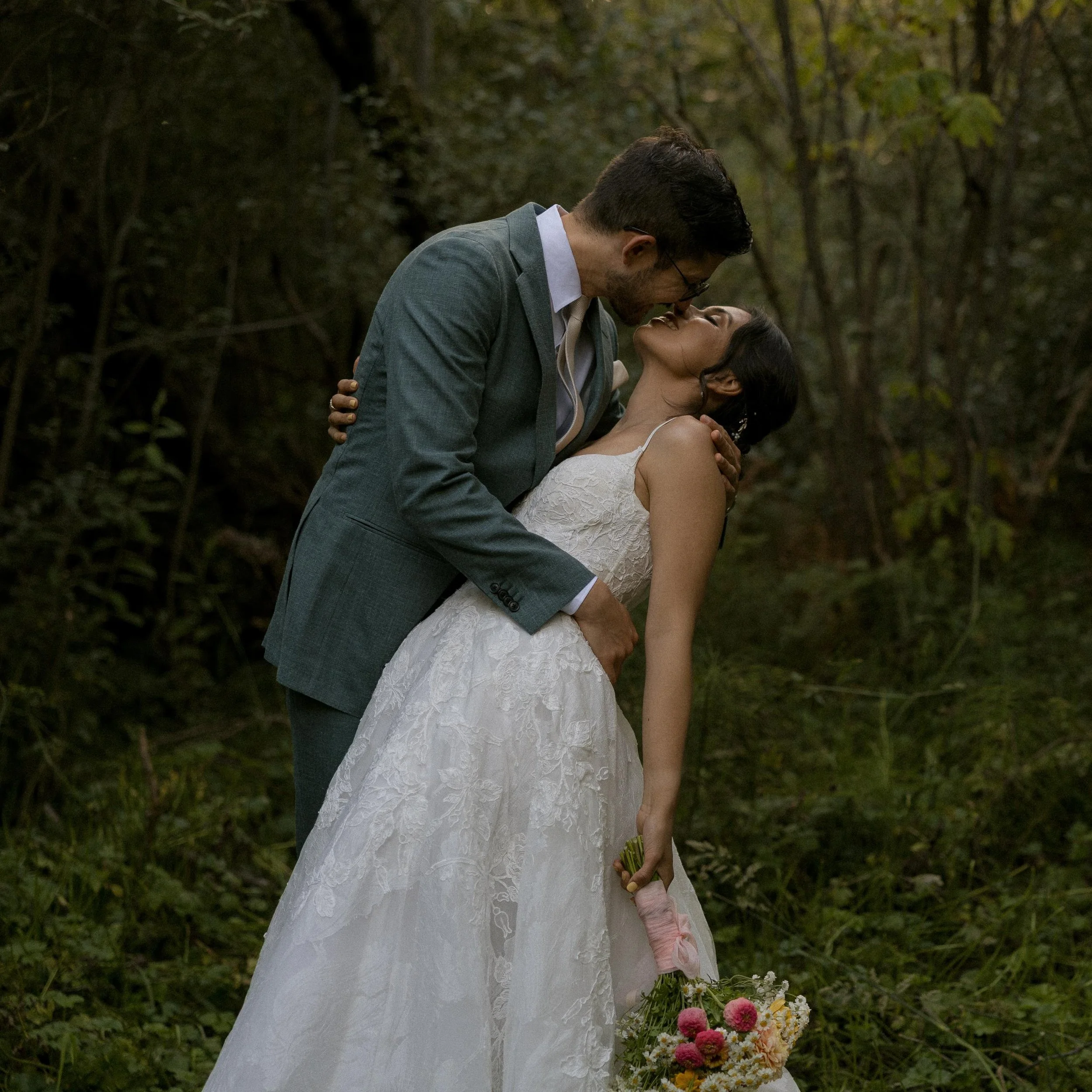 A man in a gray suit and a woman in a white lace wedding dress are kissing outdoors in a wooded area, with the woman holding a bouquet of flowers.