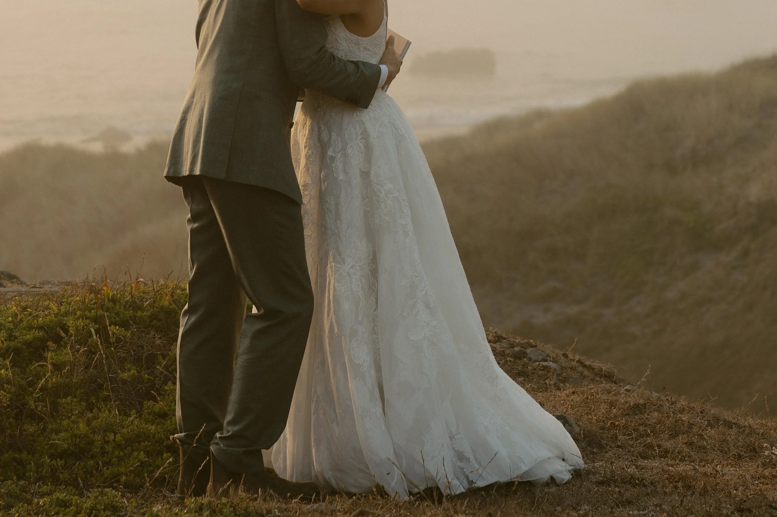 A couple dressed in wedding attire standing outdoors on a grassy hill, with a foggy or misty background.