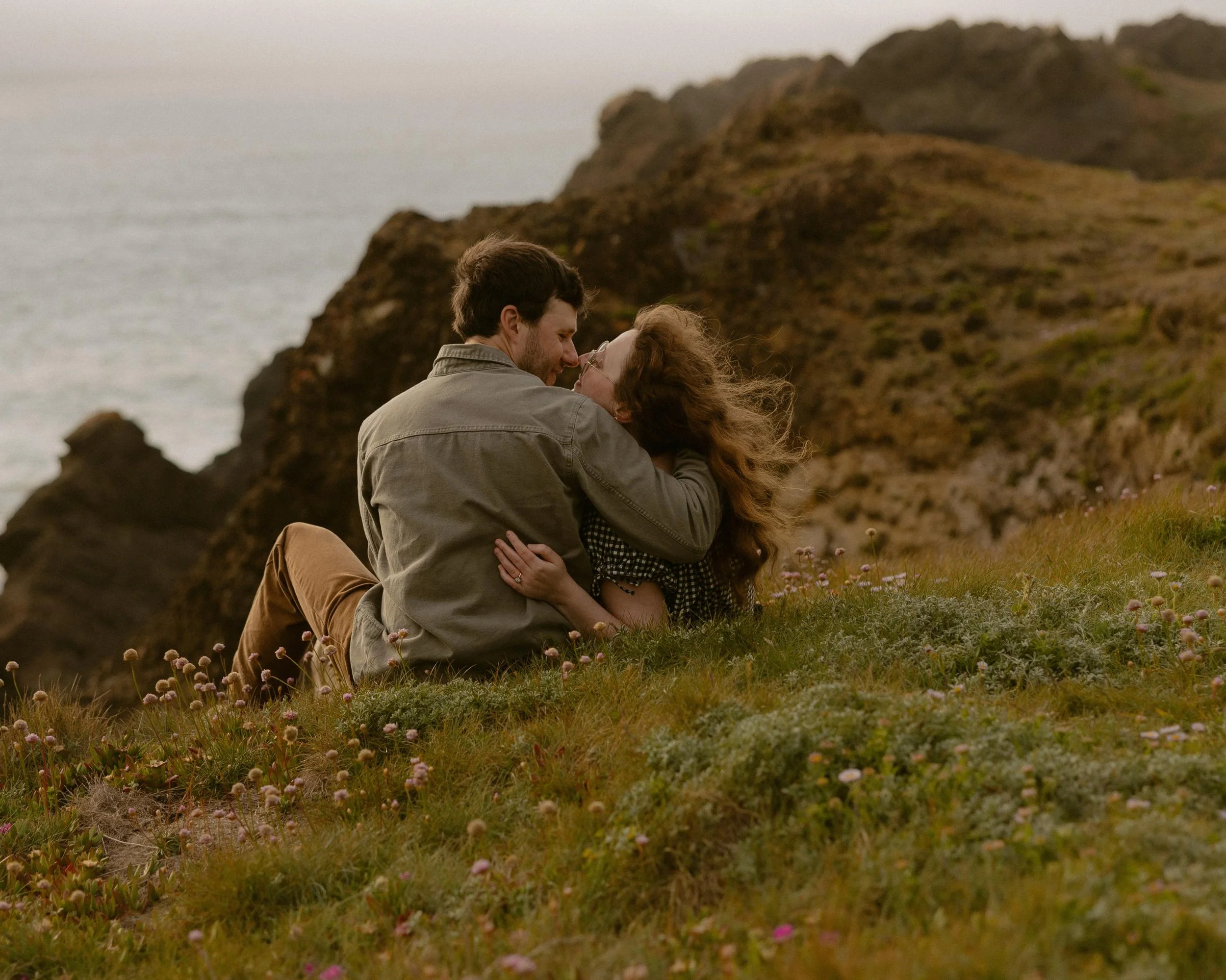 A couple sitting close together on grassy hillside with pink flowers, overlooking rocky cliffs and ocean at sunset.