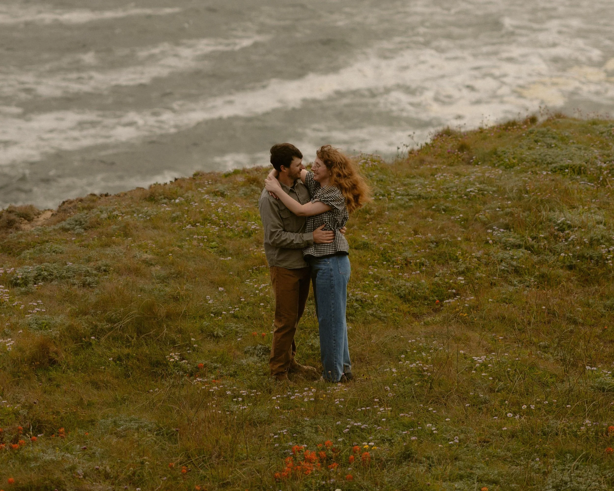 A man and woman embracing on a grassy hill near the ocean, with waves crashing in the background.