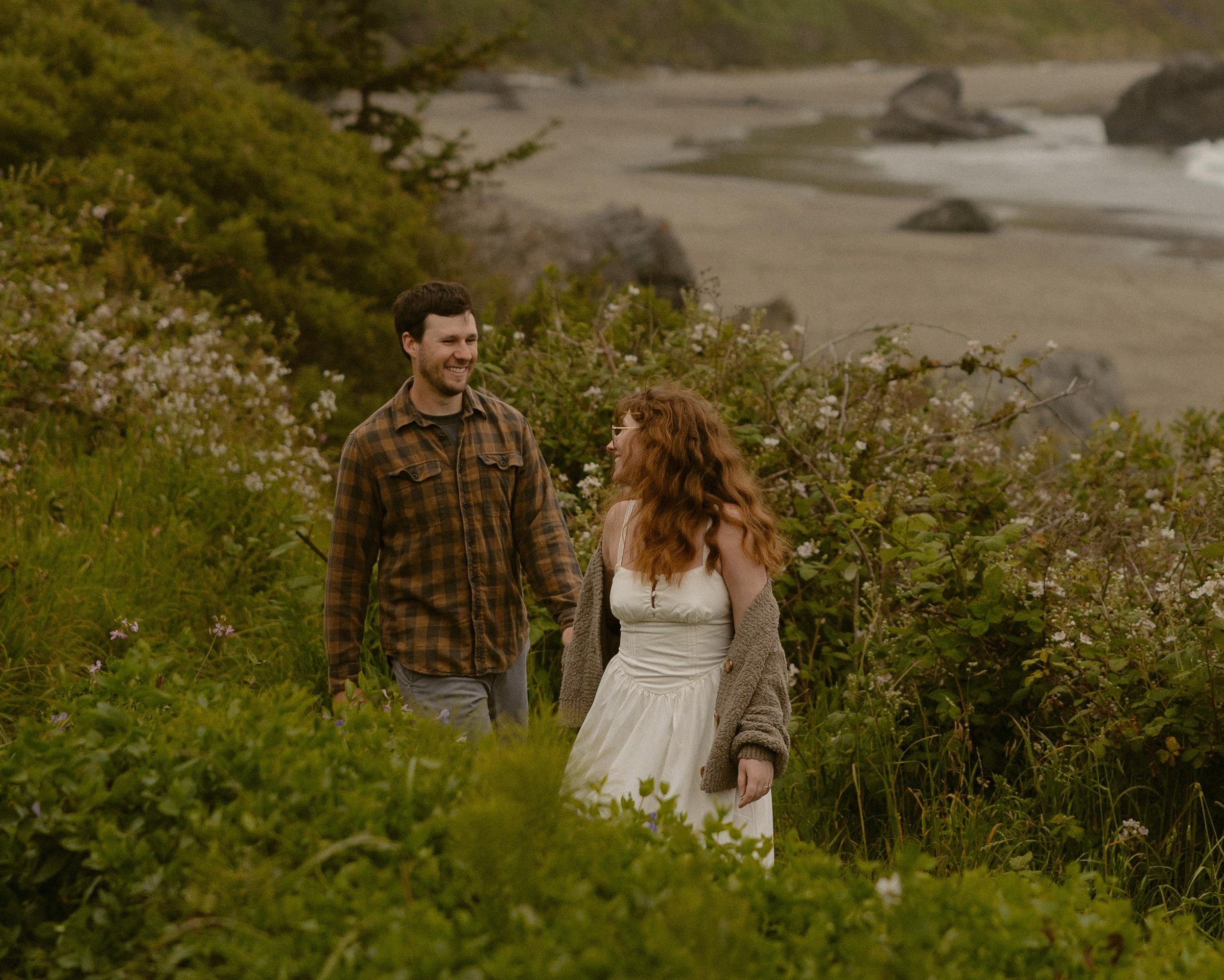 A man with dark hair and beard in a plaid shirt smiles and looks at a red-haired woman in a white dress, on a beach with green shrubbery and large rocks in the background.
