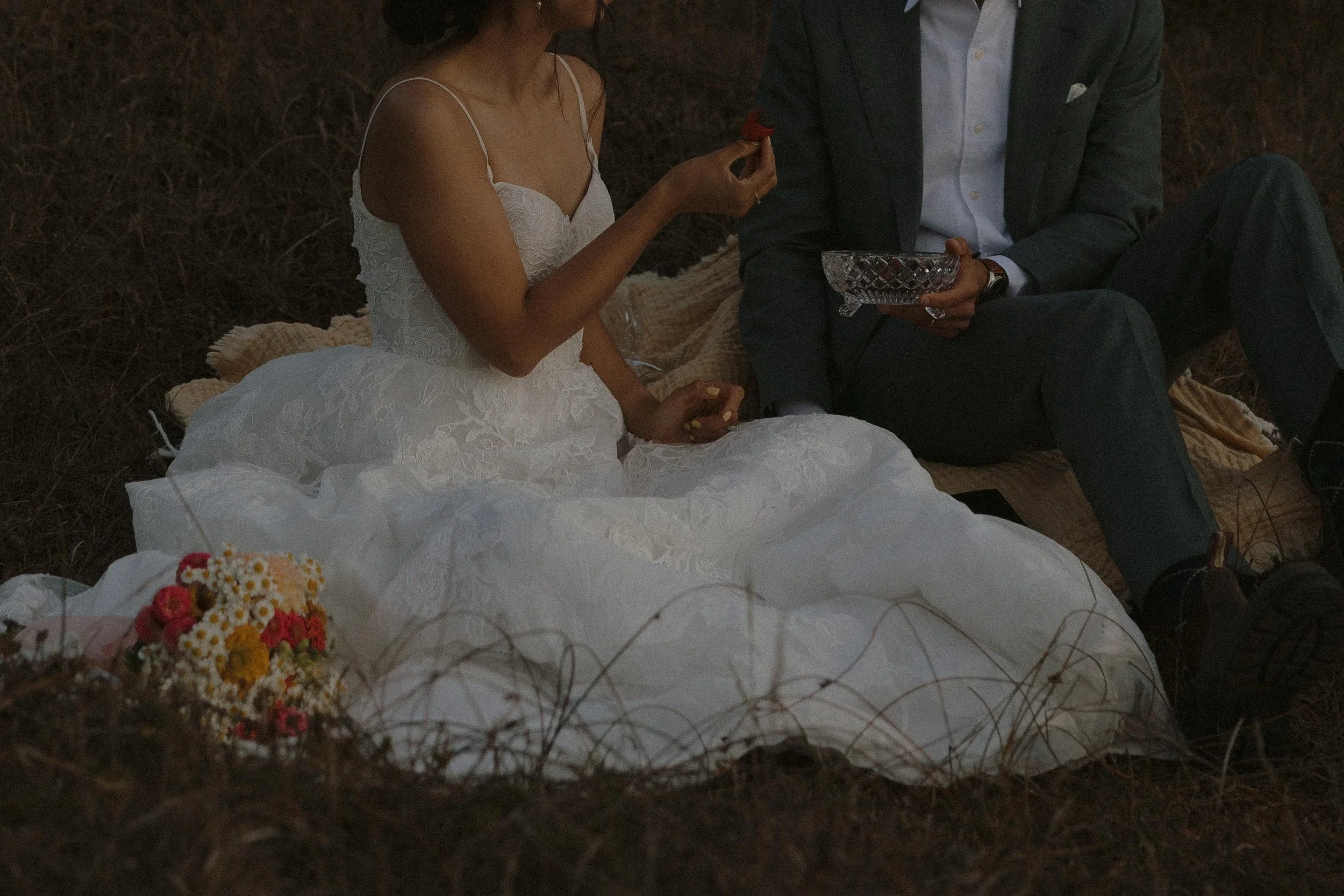 A man and a woman sitting on a blanket outdoors, with the woman in a white dress holding a red flower, and the man in a dark suit holding a glass bowl.