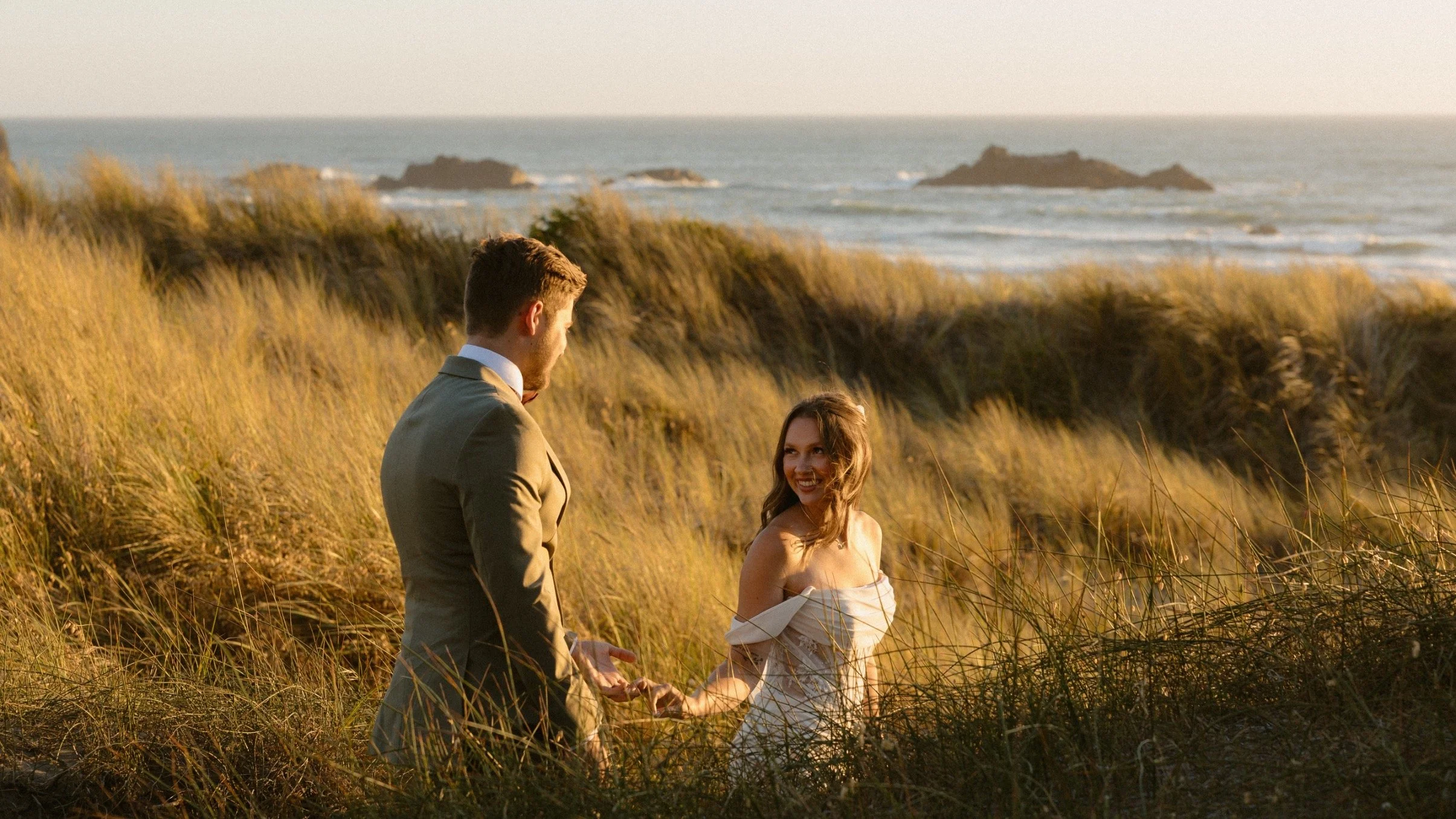 A couple standing on a grassy beach at sunset, holding hands and facing each other with the ocean and rocks in the background.