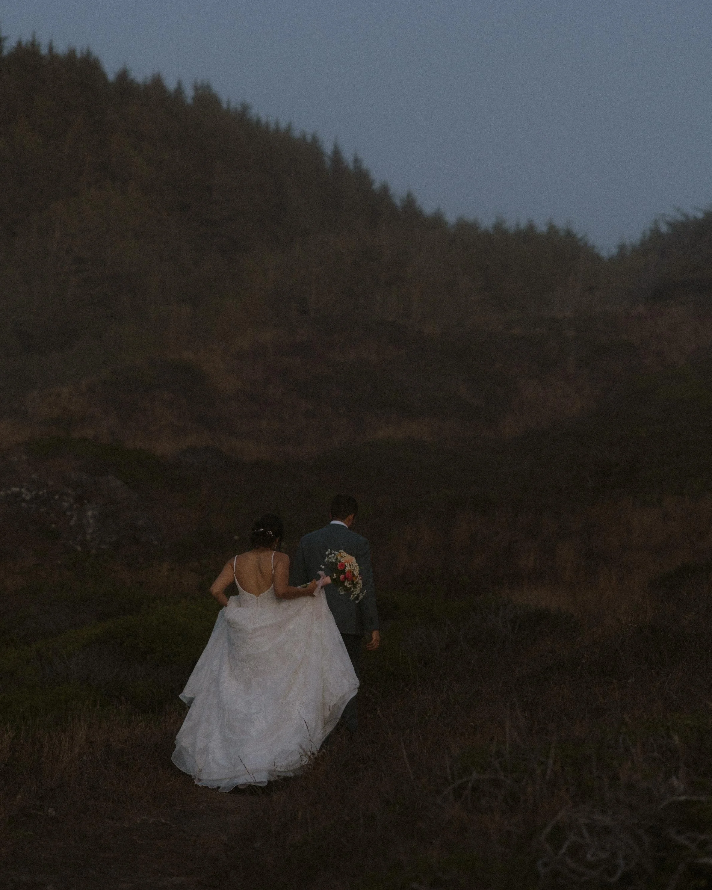 A bride and groom walking together outdoors during dusk, with the bride wearing a white gown and the groom in a dark suit, holding a bouquet of flowers.