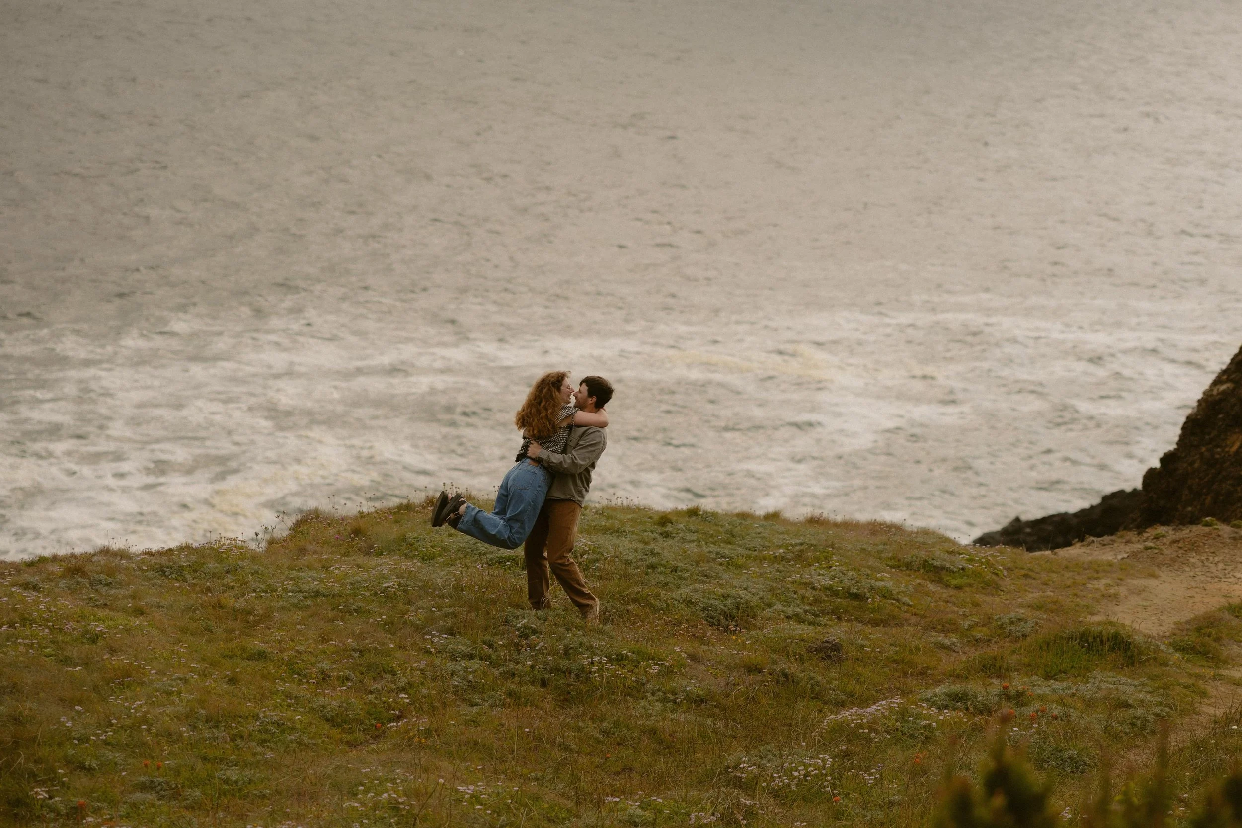 A couple on the grassy coastline during sunset, with the man lifting the woman while they face each other, with the ocean in the background.