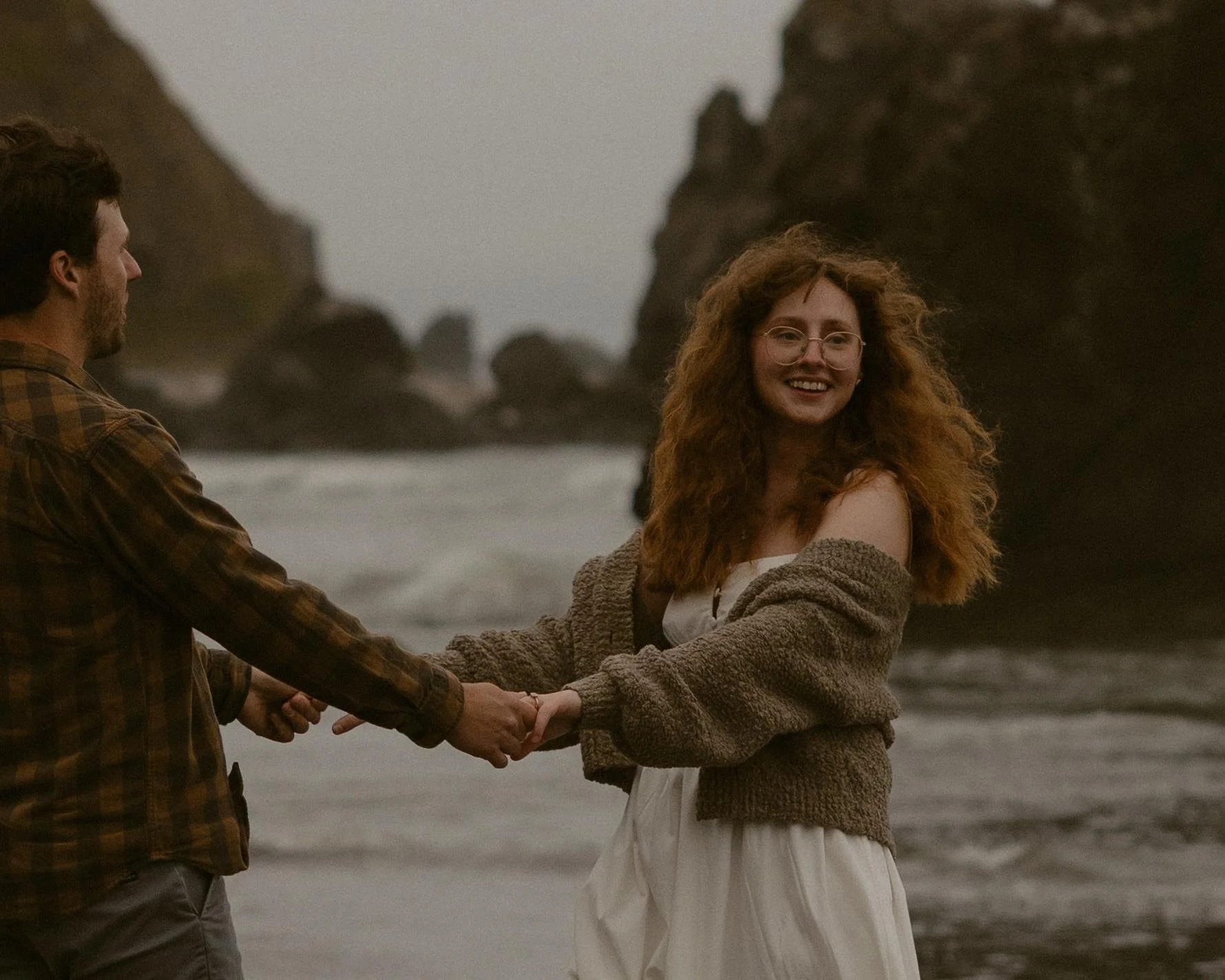 A man and a woman holding hands on a beach with large rocks in the background, overcast sky, woman smiling, wearing glasses, woman has curly red hair, man has dark hair and a beard, they are dressed casually.