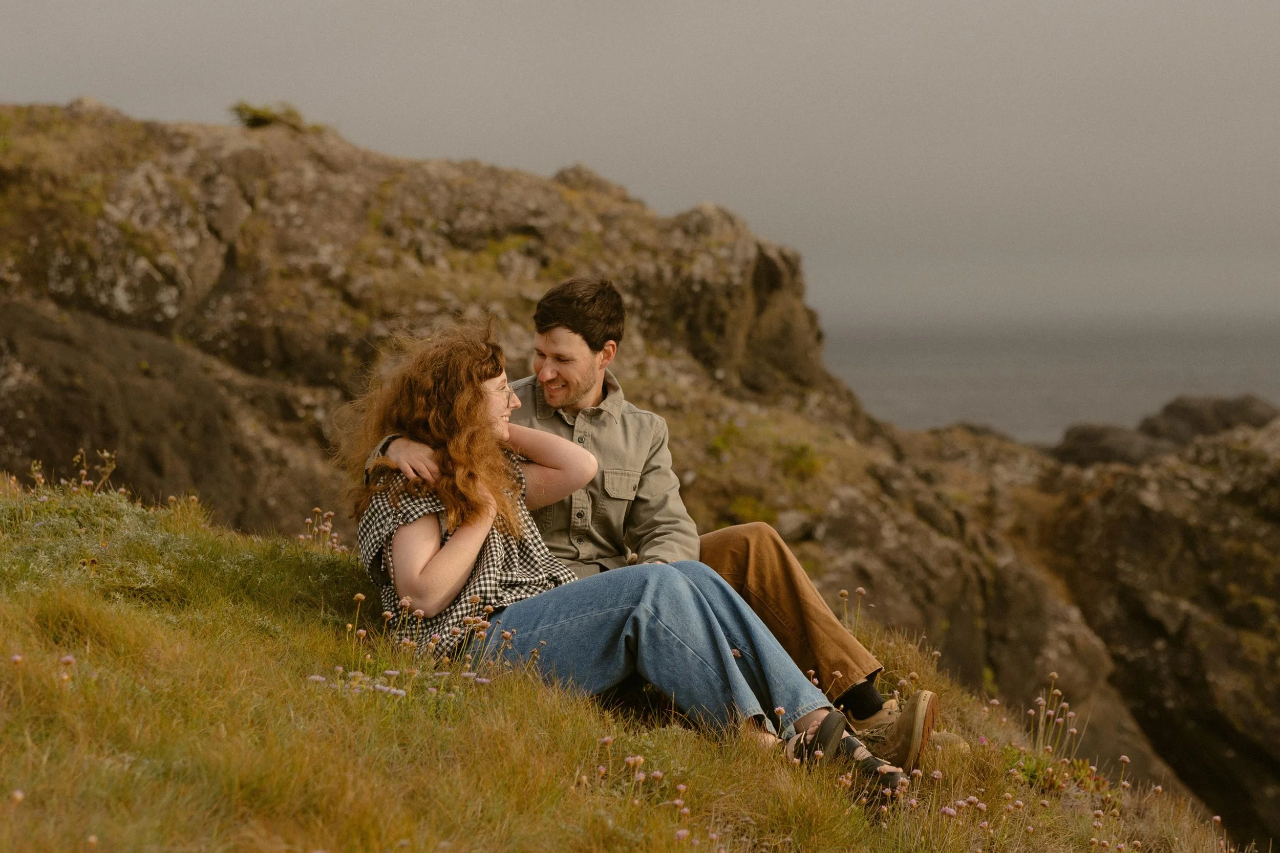 A man and woman sitting on a grassy hillside near the ocean, smiling and looking at each other, during sunset or early evening.