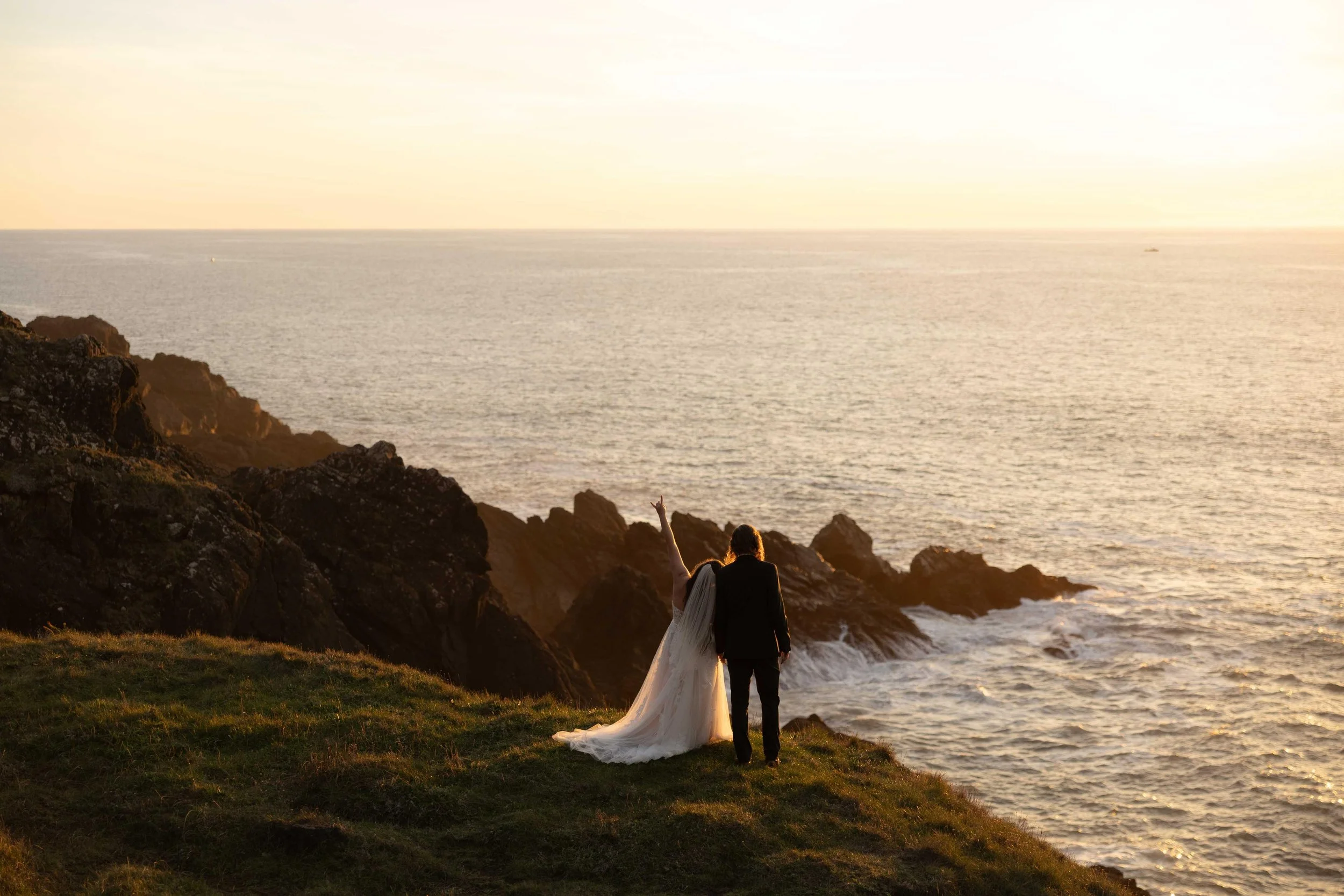 A couple dressed in wedding attire sitting on a grassy cliffside overlooking an ocean at sunset, with rocky formations on the shoreline.