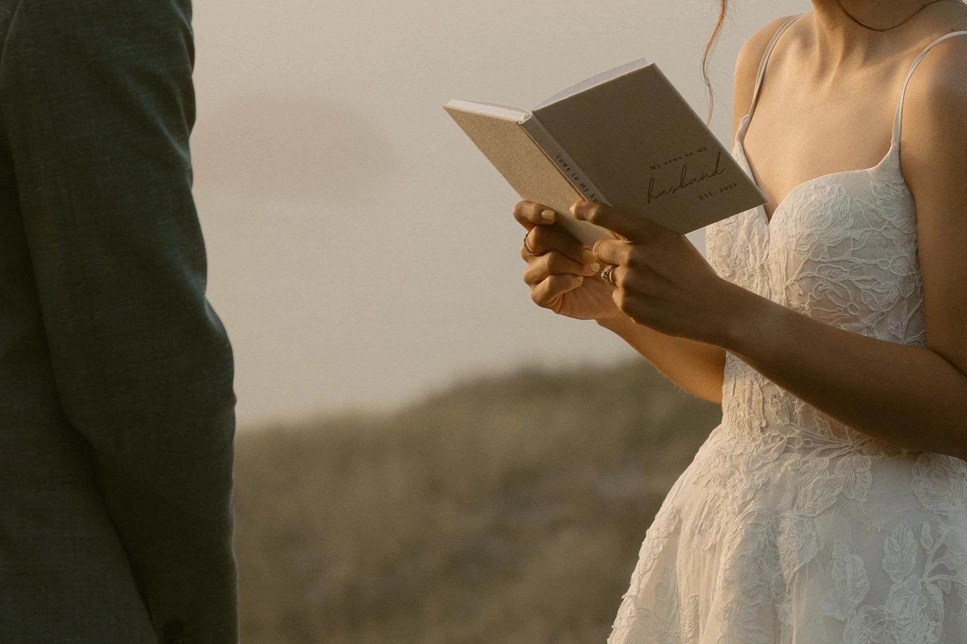 A woman in a white lace dress holding a book during a wedding ceremony, standing next to a man in black clothing.