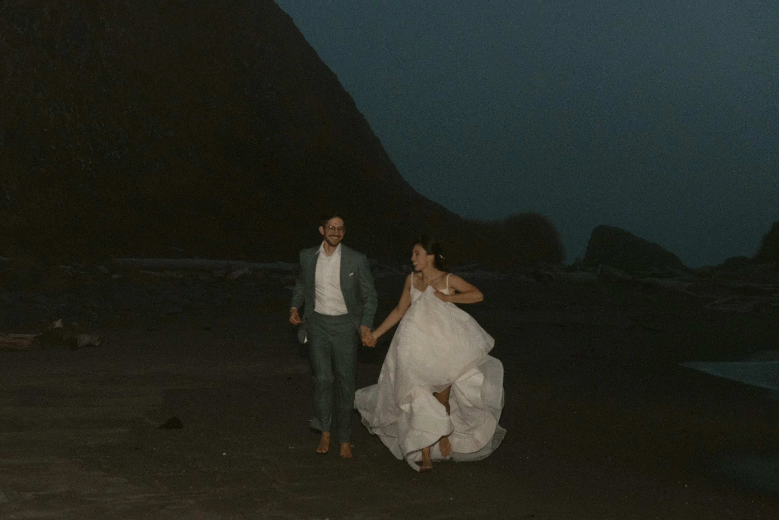 A happy couple in wedding attire walking on a beach at night, held by hands, with tall dark cliffs in the background.