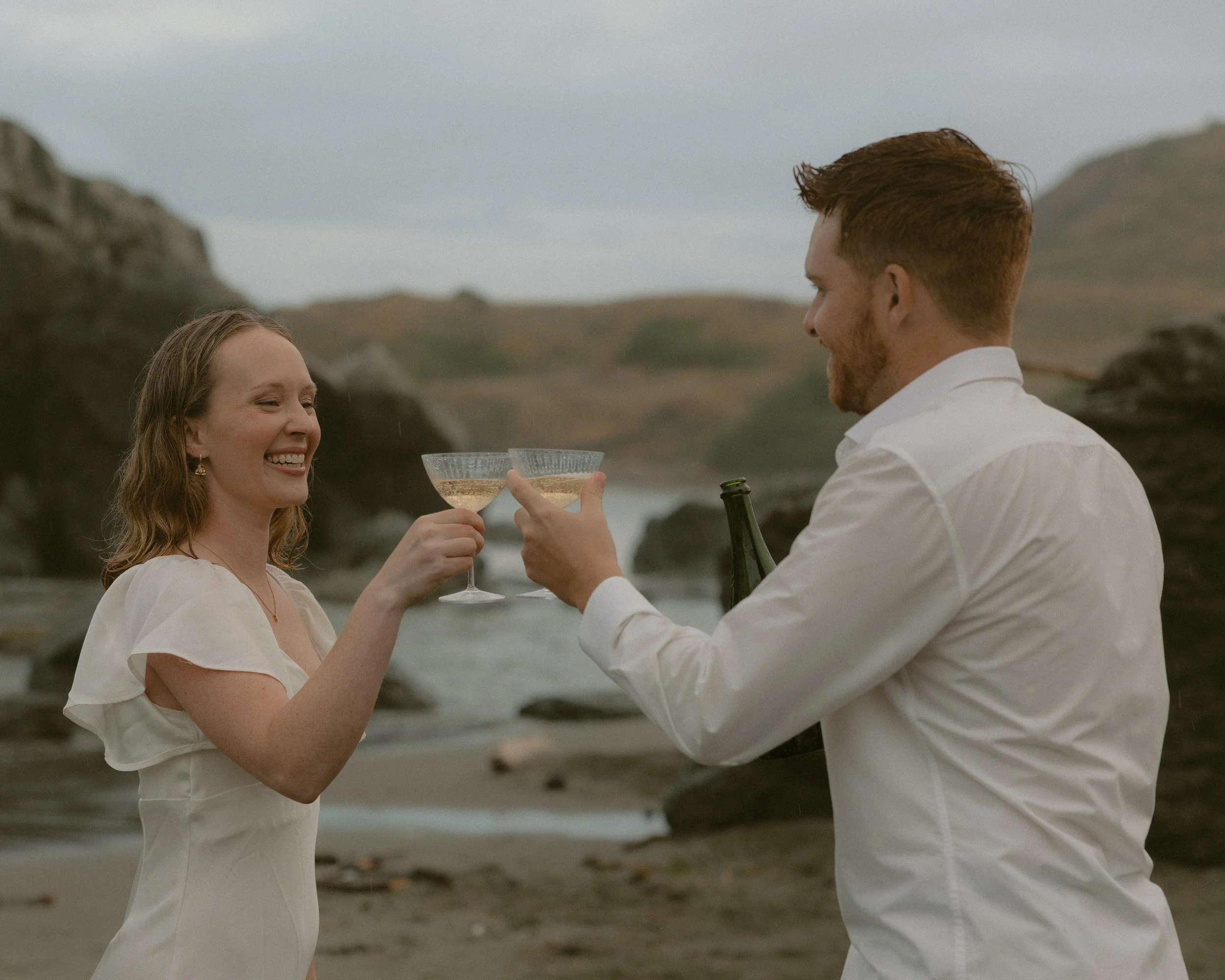 A woman and a man in white clothing are smiling and toasting with champagne glasses on a beach. The woman has red hair and is wearing a white dress, while the man has red hair and a beard. They are outdoors with rocky terrain and water in the backgro