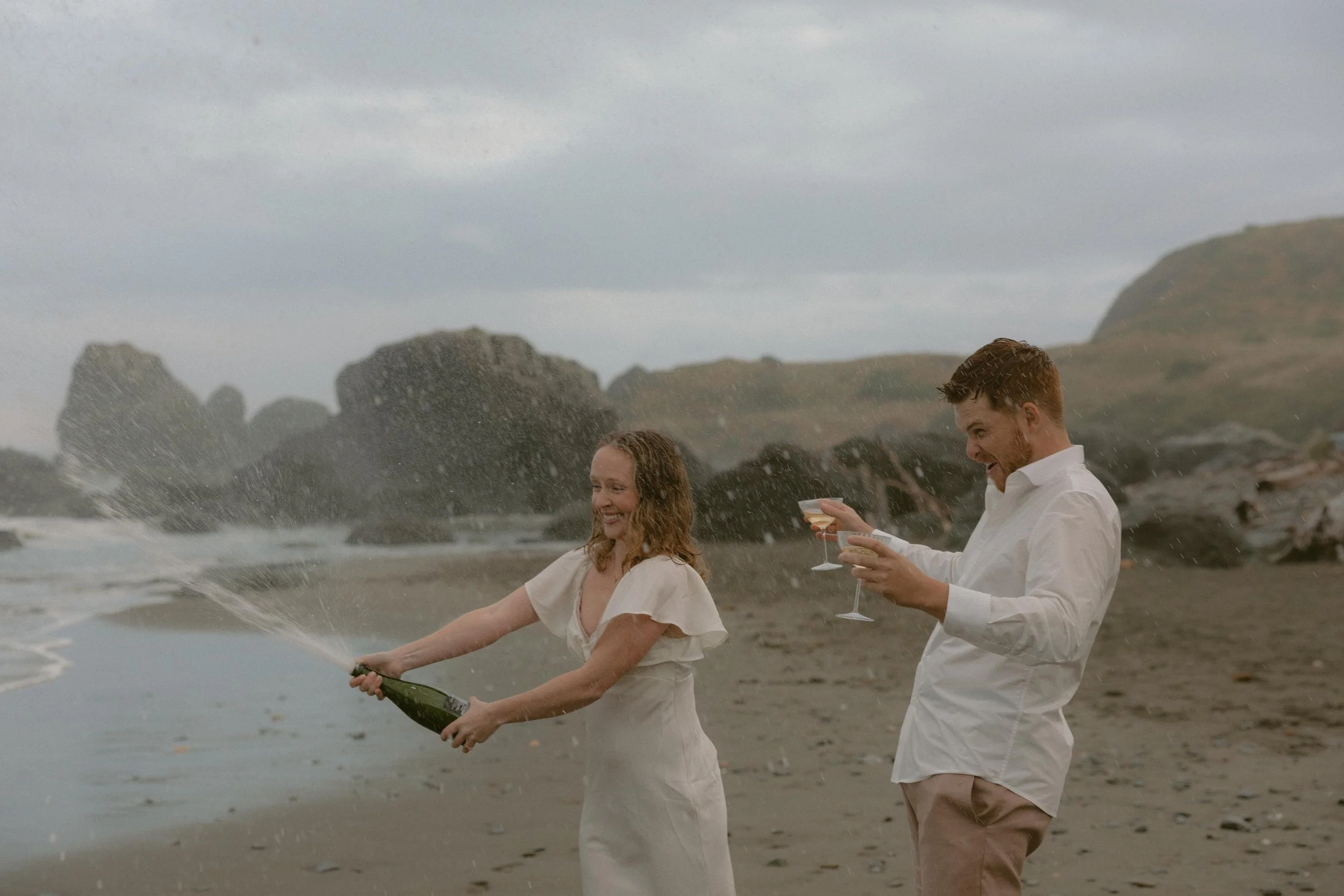 A man and woman celebrating on a beach, opening a bottle of champagne and spraying it as they smile, with rocky cliffs and cloudy sky in the background.