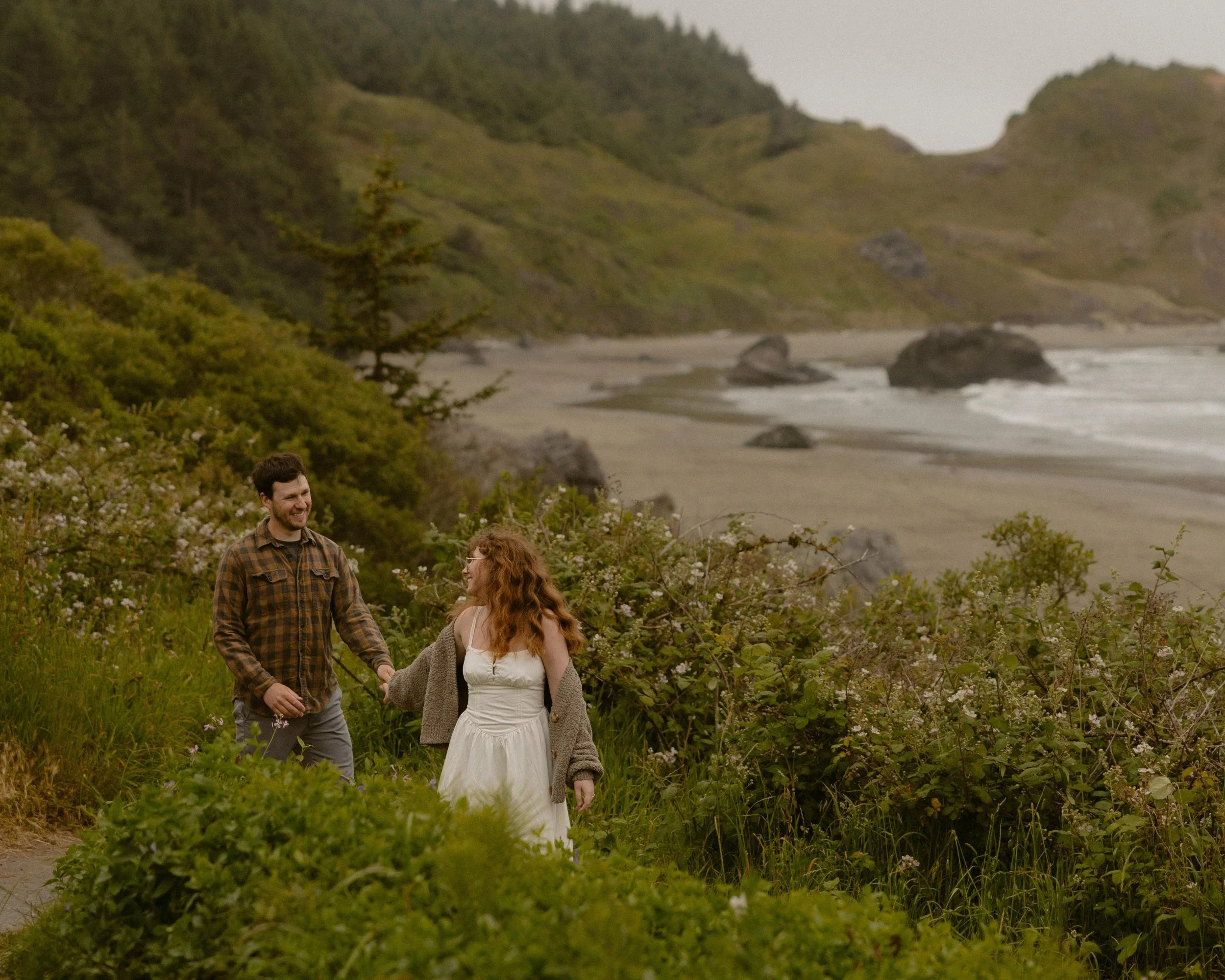 Three people are walking through a lush green field filled with low plants and shrubs, near a beach with rocks and ocean waves. In the background, green hills and cliffs extend along the coastline under an overcast sky. Two women and one man are smil