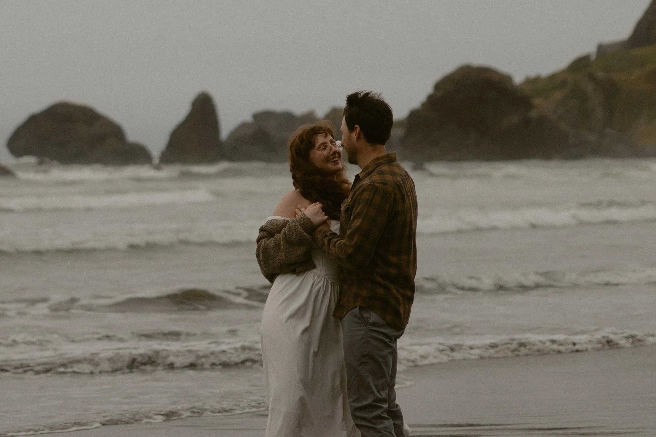 A couple standing on the beach, facing each other and smiling, with rocky formations in the background.