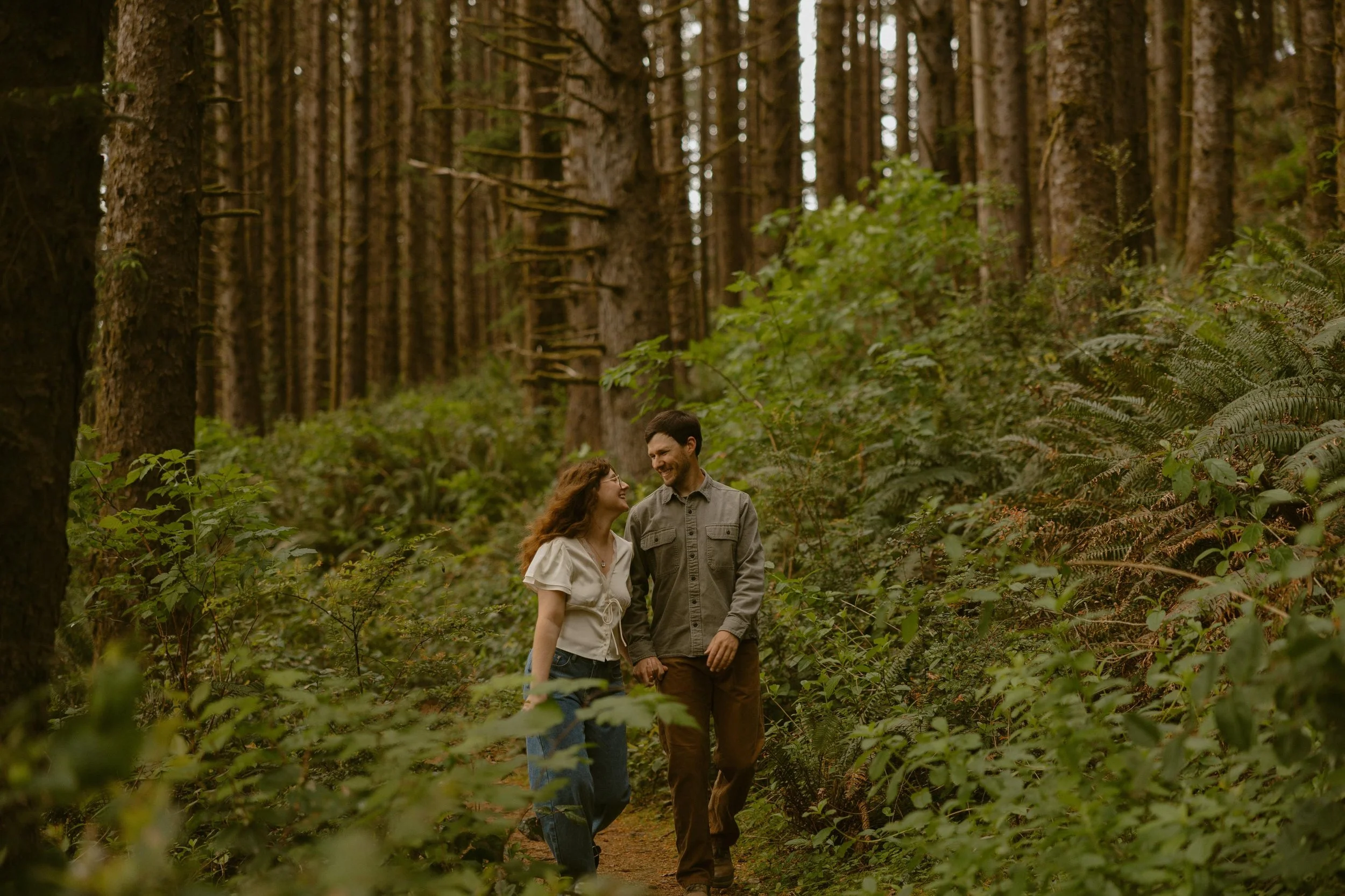 A couple walking in a lush green forest with tall trees and ferns, smiling and looking at each other.