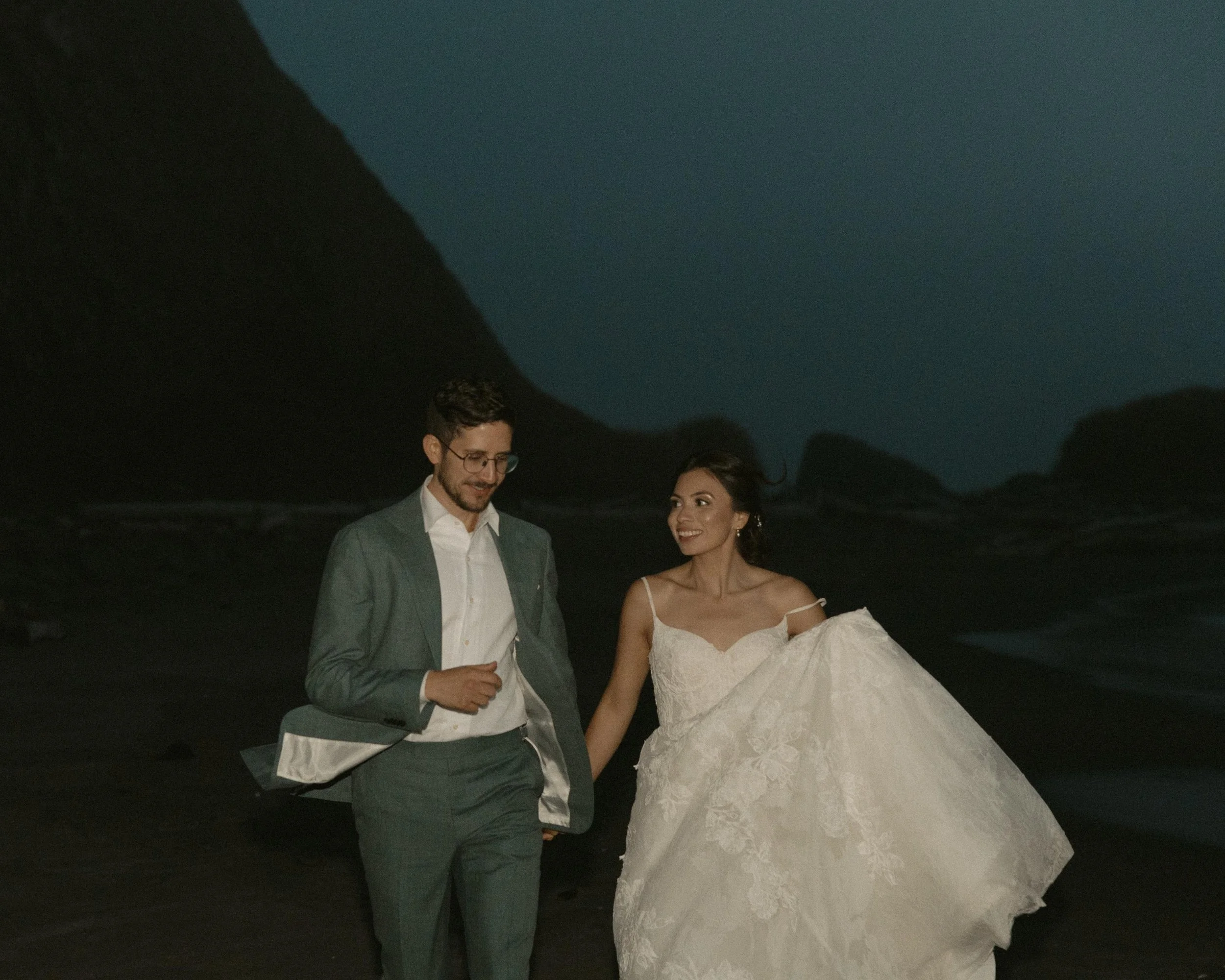 A man and woman in wedding attire walking outdoors at night, holding hands with mountains and a dark sky in the background.