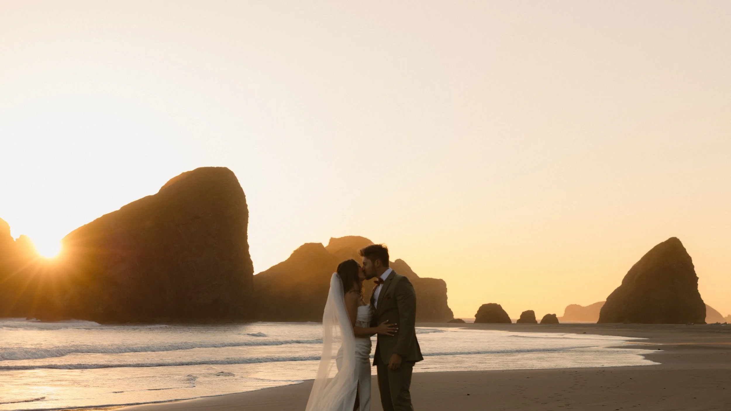 A bride and groom standing close together on the beach during sunset, kissing with large rock formations and the ocean in the background.