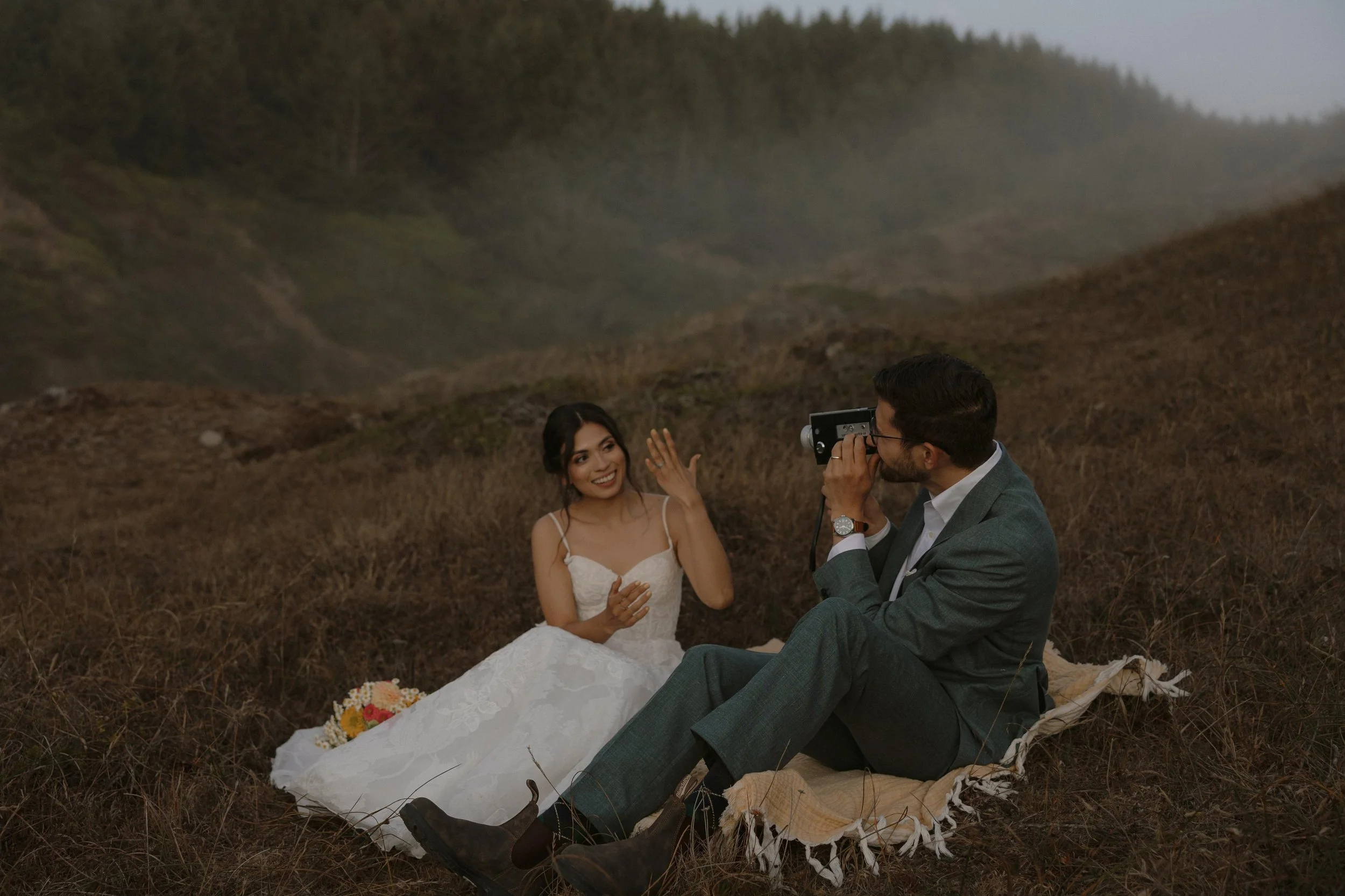 A man and woman in wedding attire sitting on the ground in a natural outdoor setting with grass and hills, engaged in a photo shoot.