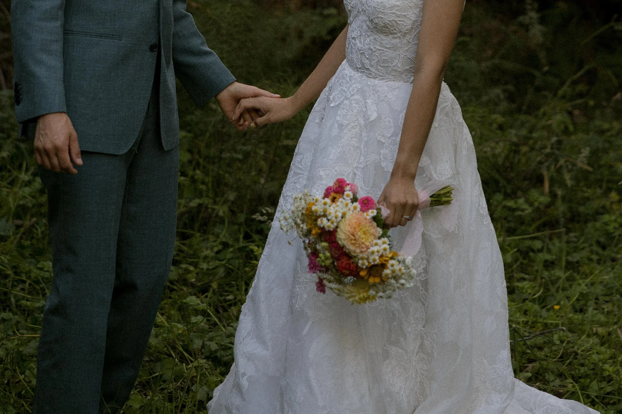 A couple holding hands during a wedding ceremony outdoors. The bride is holding a bouquet of flowers, and both are dressed in formal wedding attire.