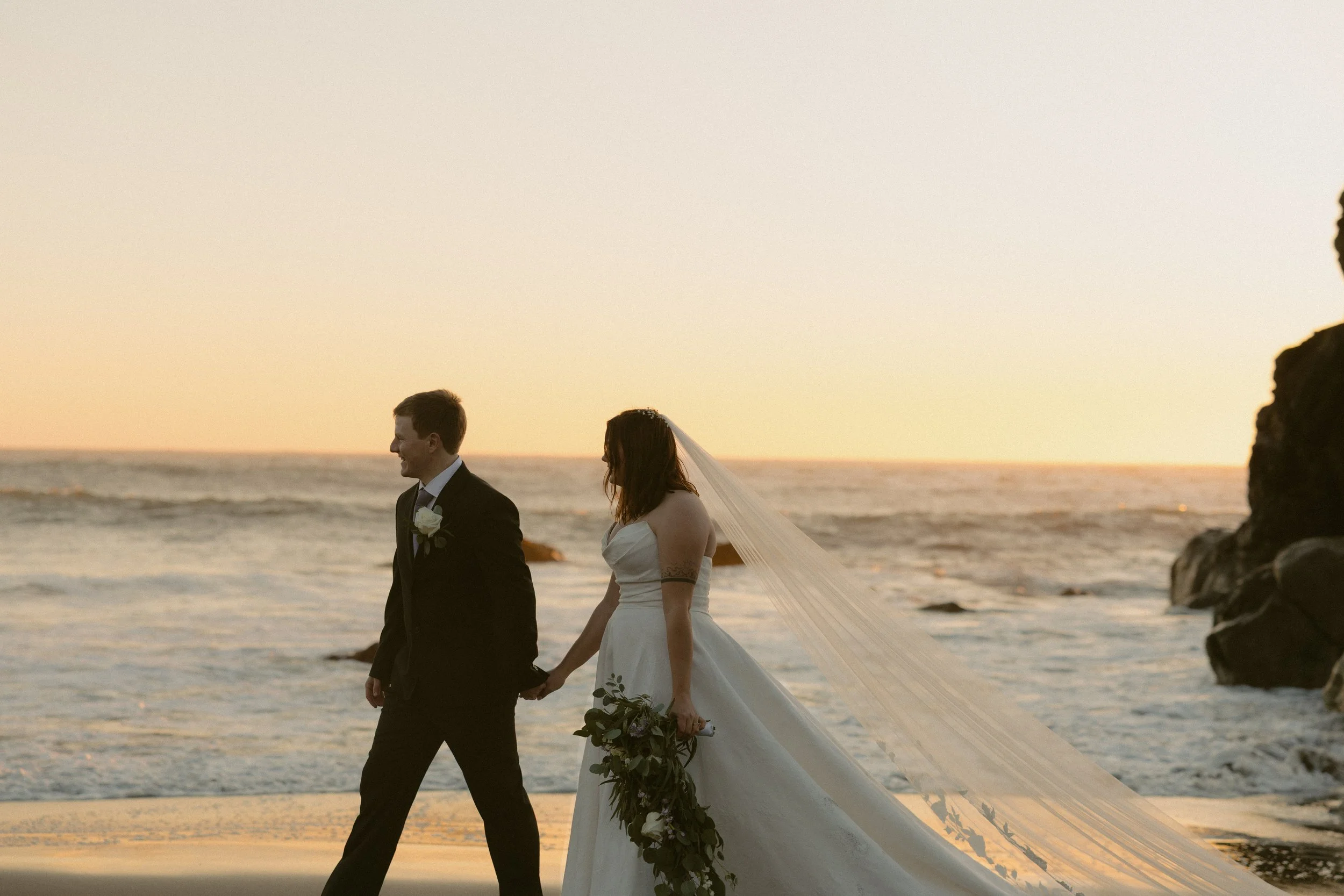 A bride and groom walking hand in hand on a beach at sunset, dressed in wedding attire, with the bride holding a bouquet of greenery and flowers.