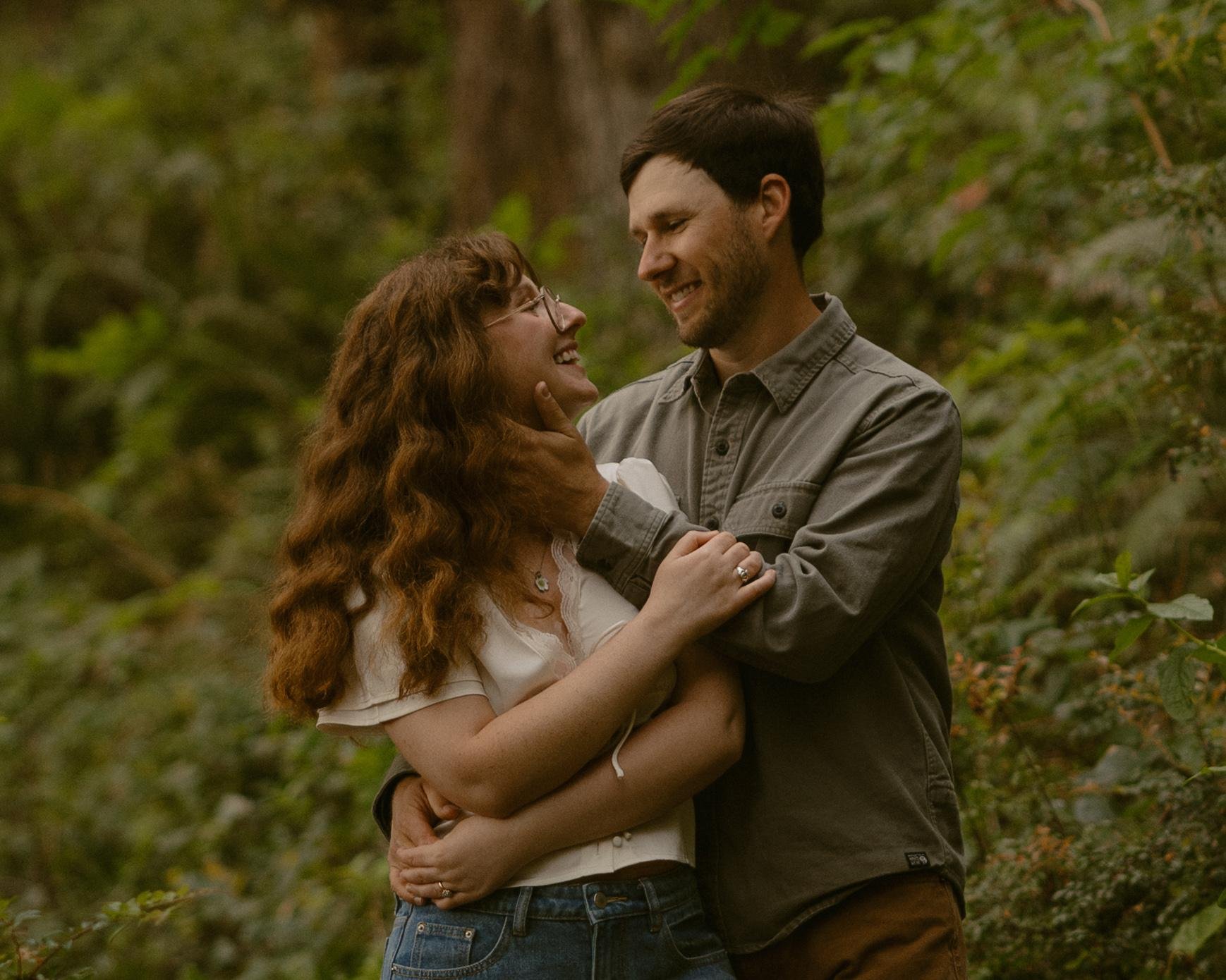 A couple sharing an intimate moment outdoors surrounded by greenery, with the man gently holding the woman's face and the woman smiling up at him.