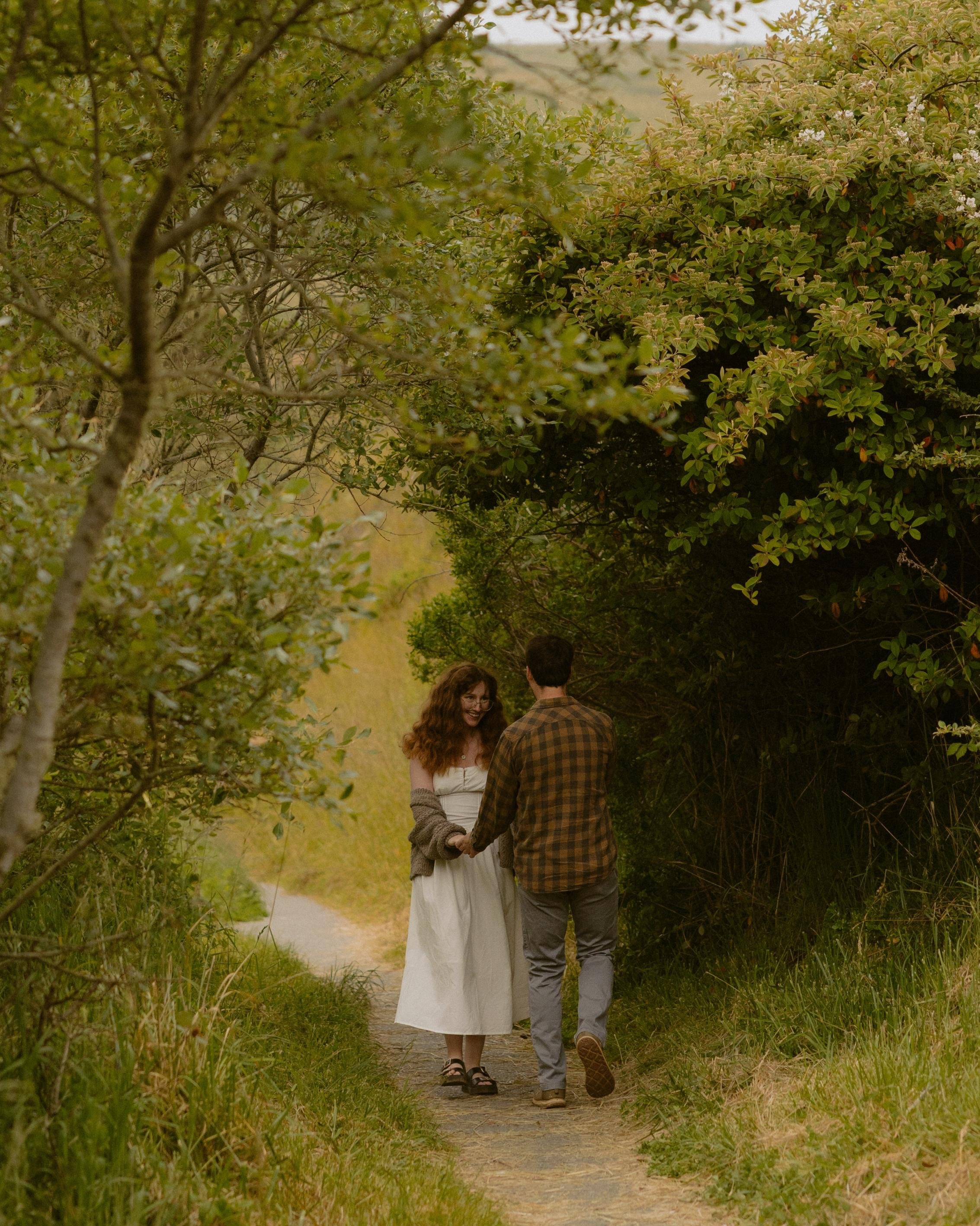 A young couple holding hands and walking along a dirt path through a lush, green, natural setting with trees and bushes.