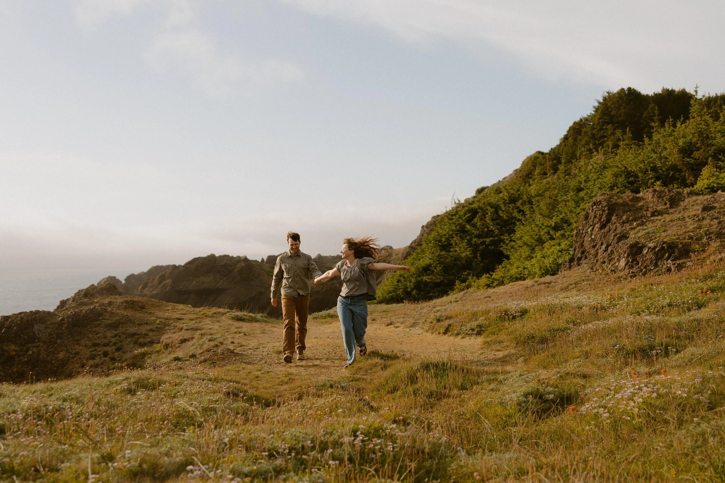 A man and woman walking together along a dirt trail on a grassy hillside with green trees and rocky formations in the background, under a partly cloudy sky.