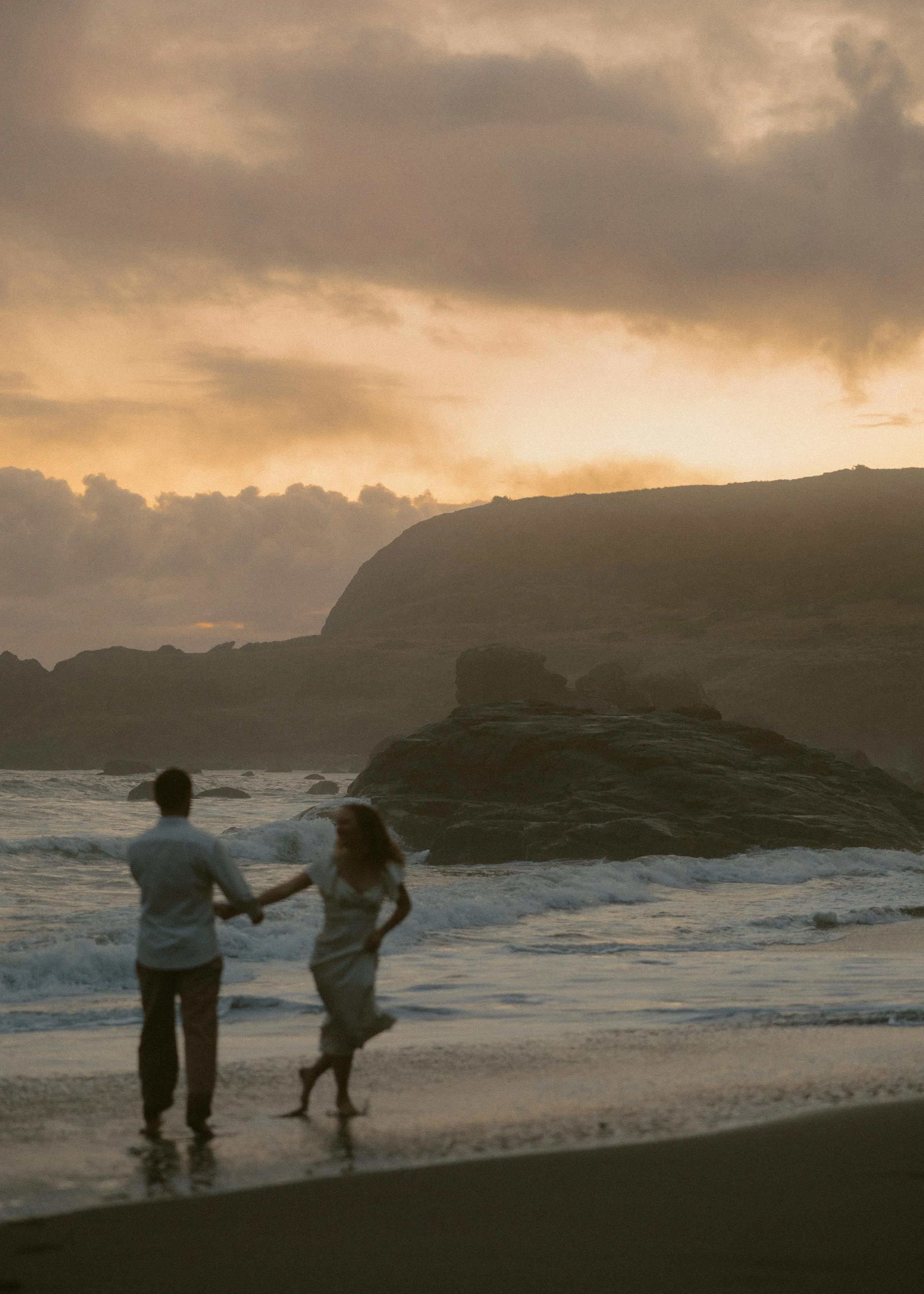 A man and woman holding hands, dancing or playing on the beach during sunset, with dark clouds, rock formations, and waves in the background.