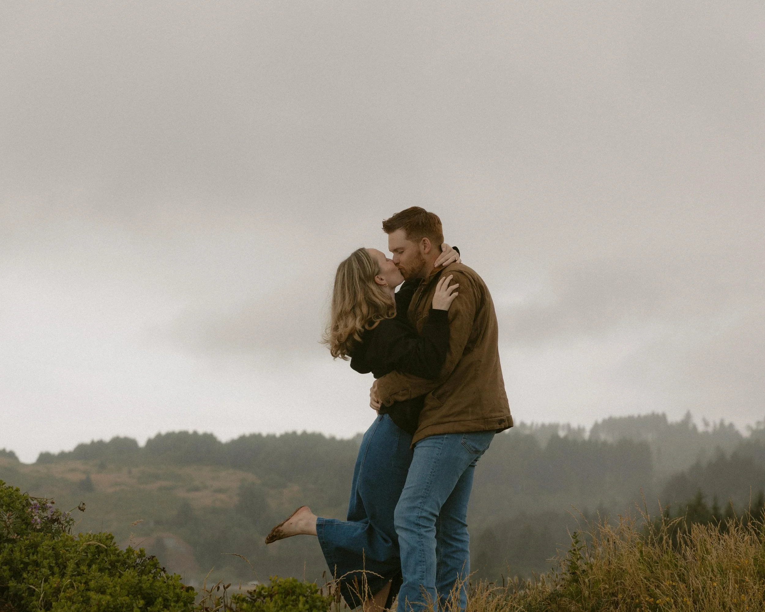 A couple embraces outdoors, with as if they are about to kiss, in a grassy field with rolling hills and a cloudy sky in the background.