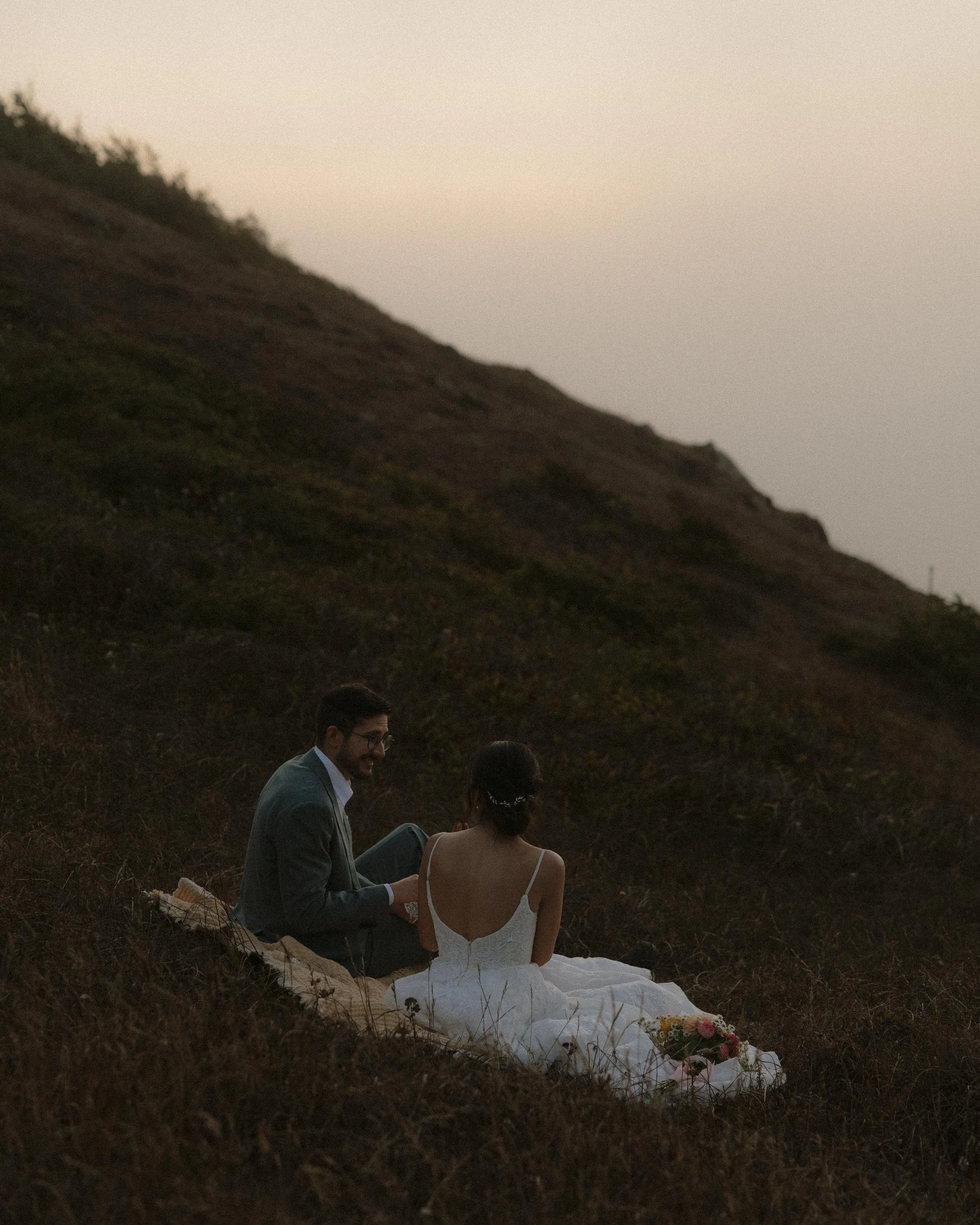 A couple dressed in formal wedding attire sitting on a grassy hillside during sunset, with the bride in a white gown and the groom in a suit, smiling and engaging in conversation.
