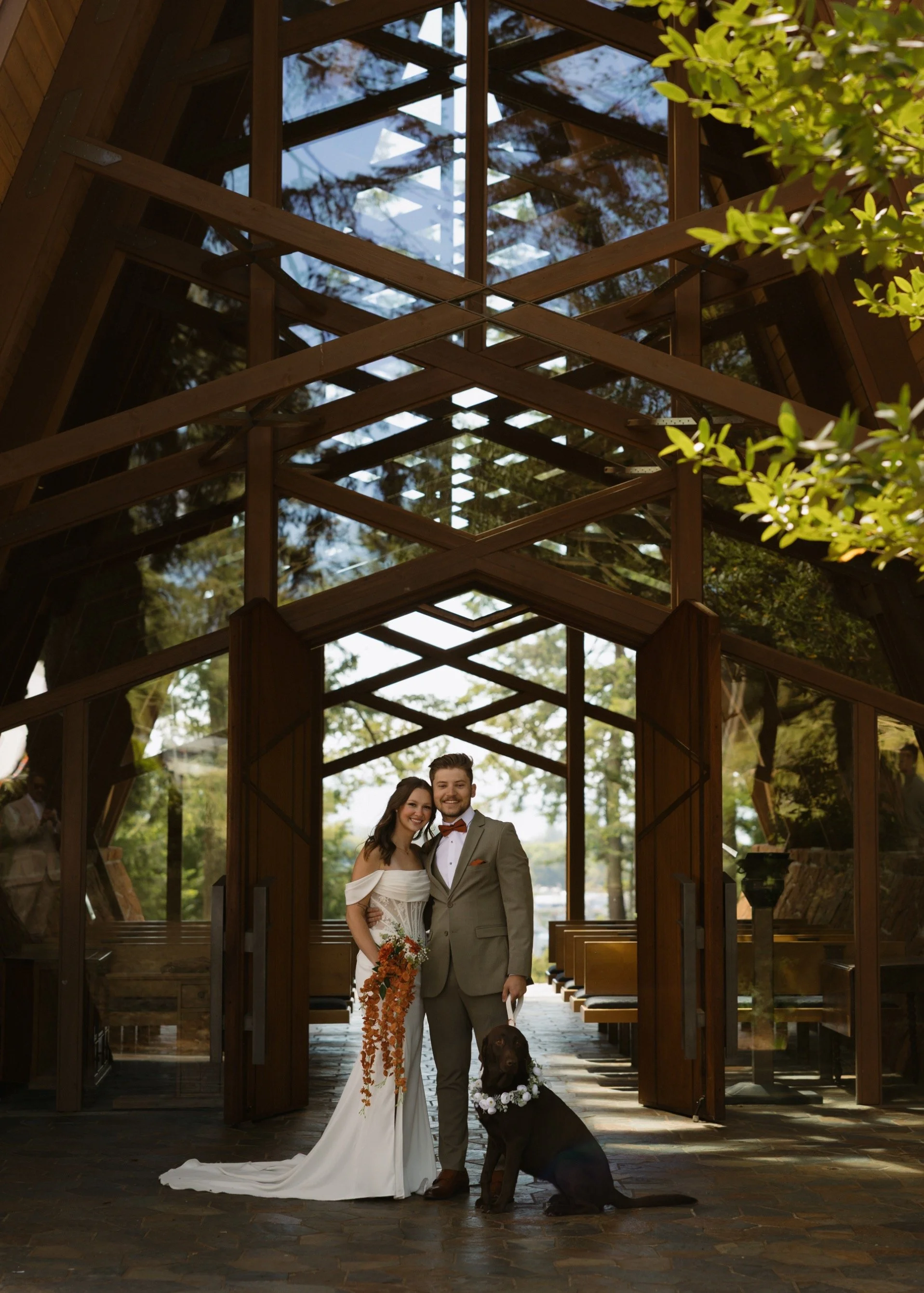 A bride and groom posing with a service dog inside a modern wooden and glass structure, with trees and greenery visible outside.