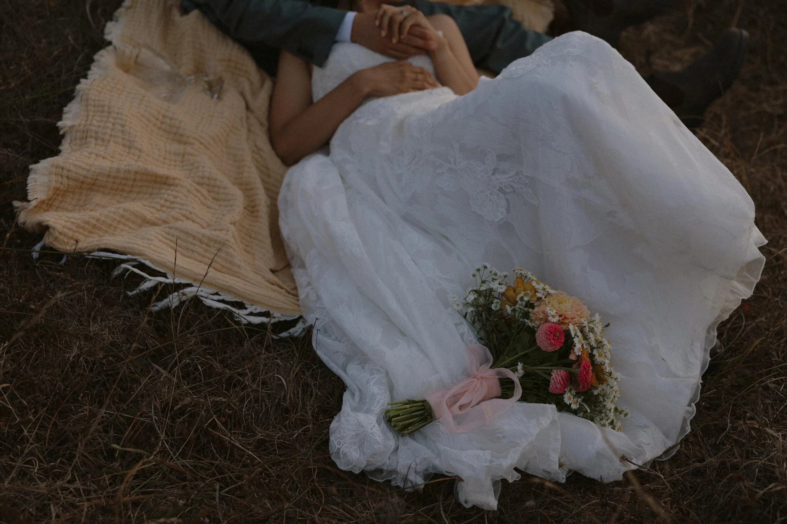 A wedding dress and bouquet of flowers lying on the ground, with a person's hand holding a child's hand in the background.