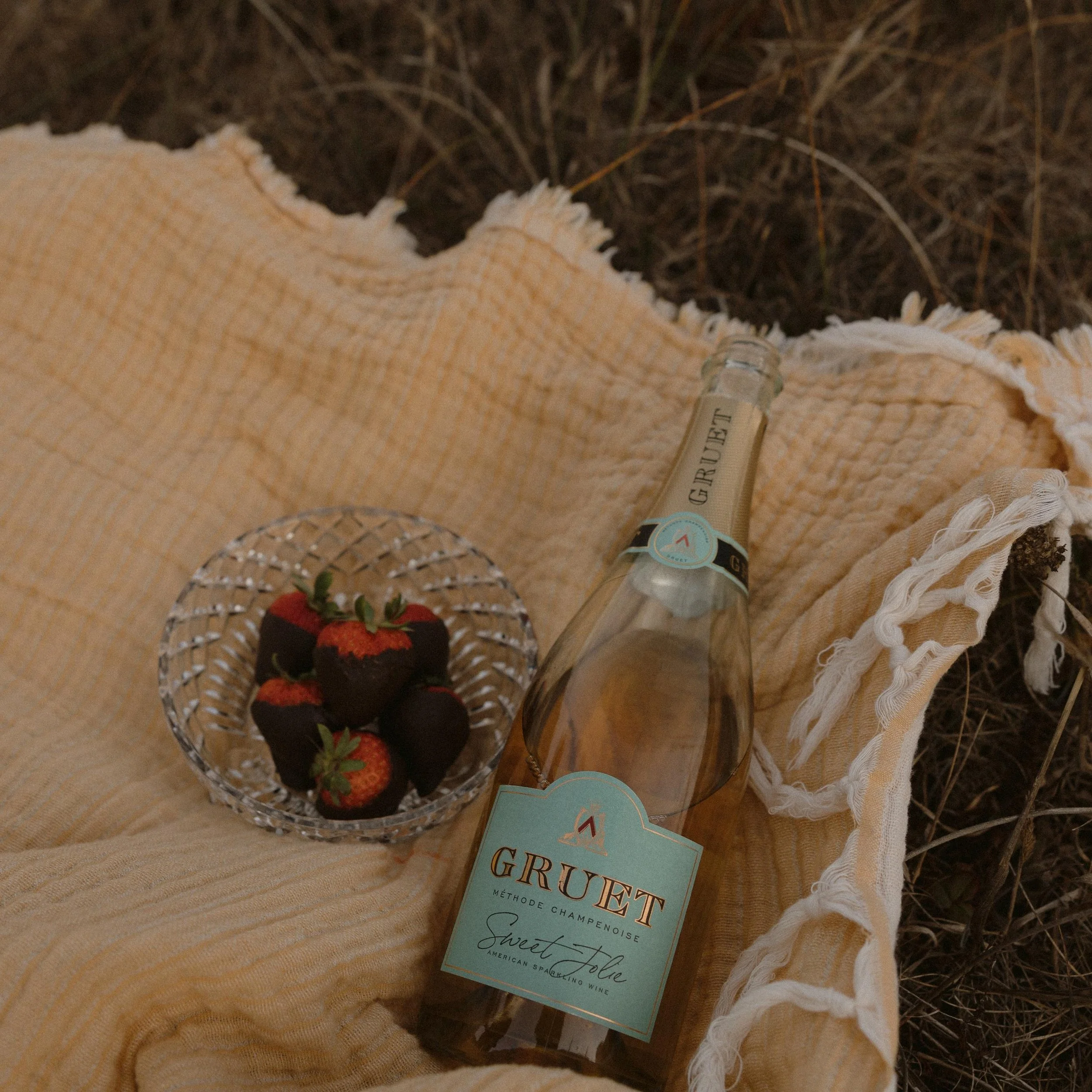A cloth with frayed edges, a glass bottle labeled "Grape" leaning on it, a small card with the words "Sweet Folk" and "Method Champagne" resting on the cloth, and a small basket of strawberries, all placed on dry grass.