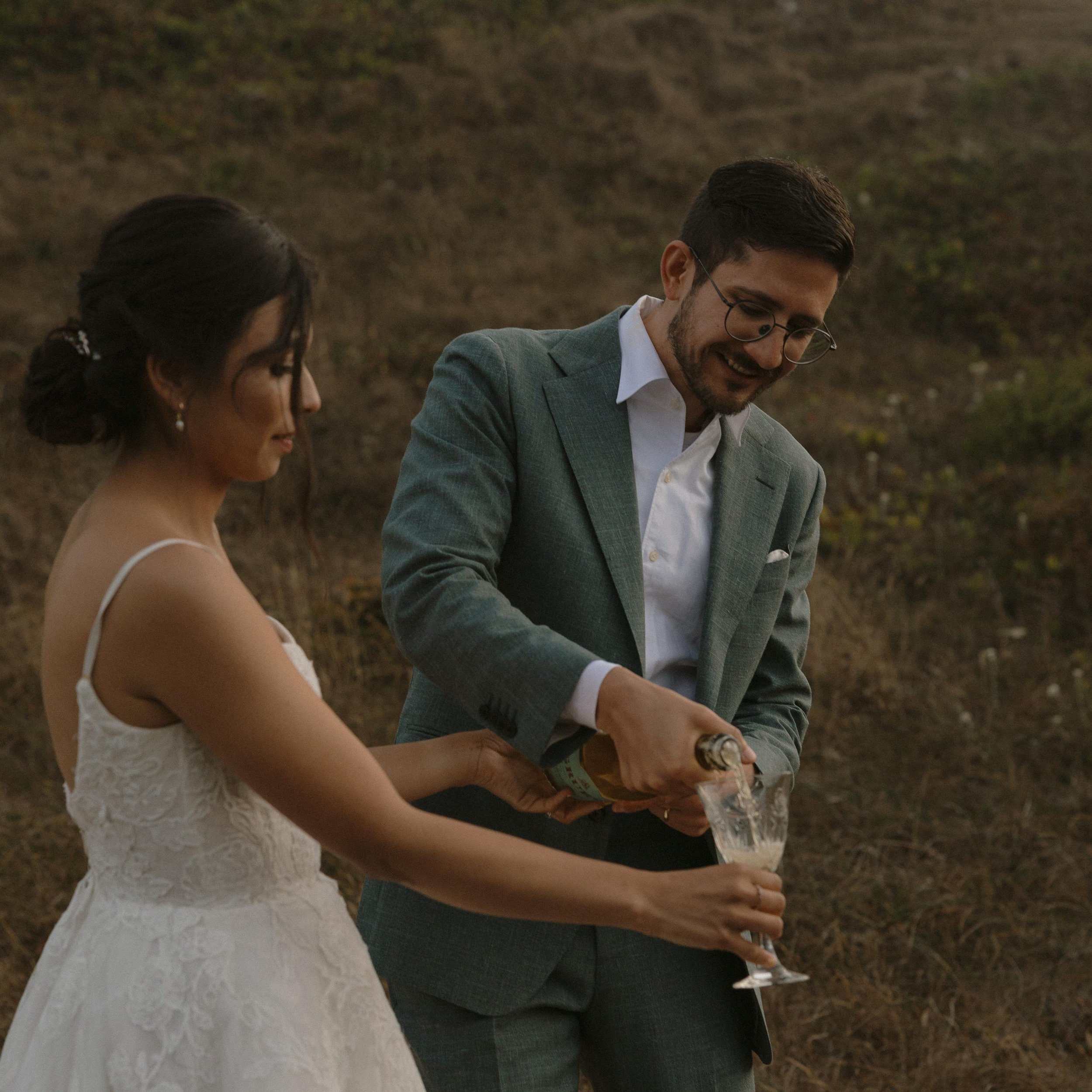 A bride and groom standing outdoors, engaged in a toast or celebration, with the bride in a white lace dress and the groom in a gray suit with glasses, holding a bottle.
