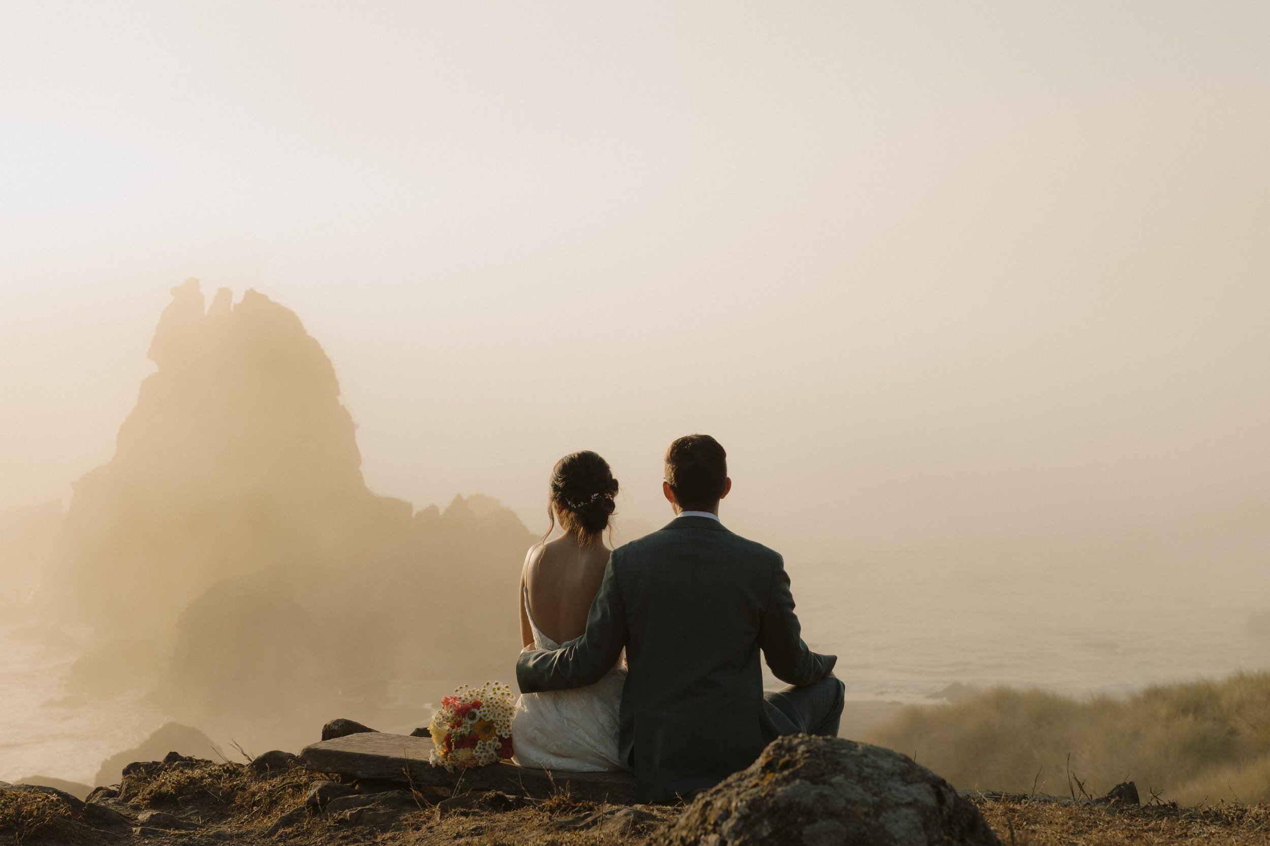 A couple, dressed in wedding attire, sitting close together outdoors during sunset or sunrise, with the bride holding a bouquet, facing a scenic mountain view.