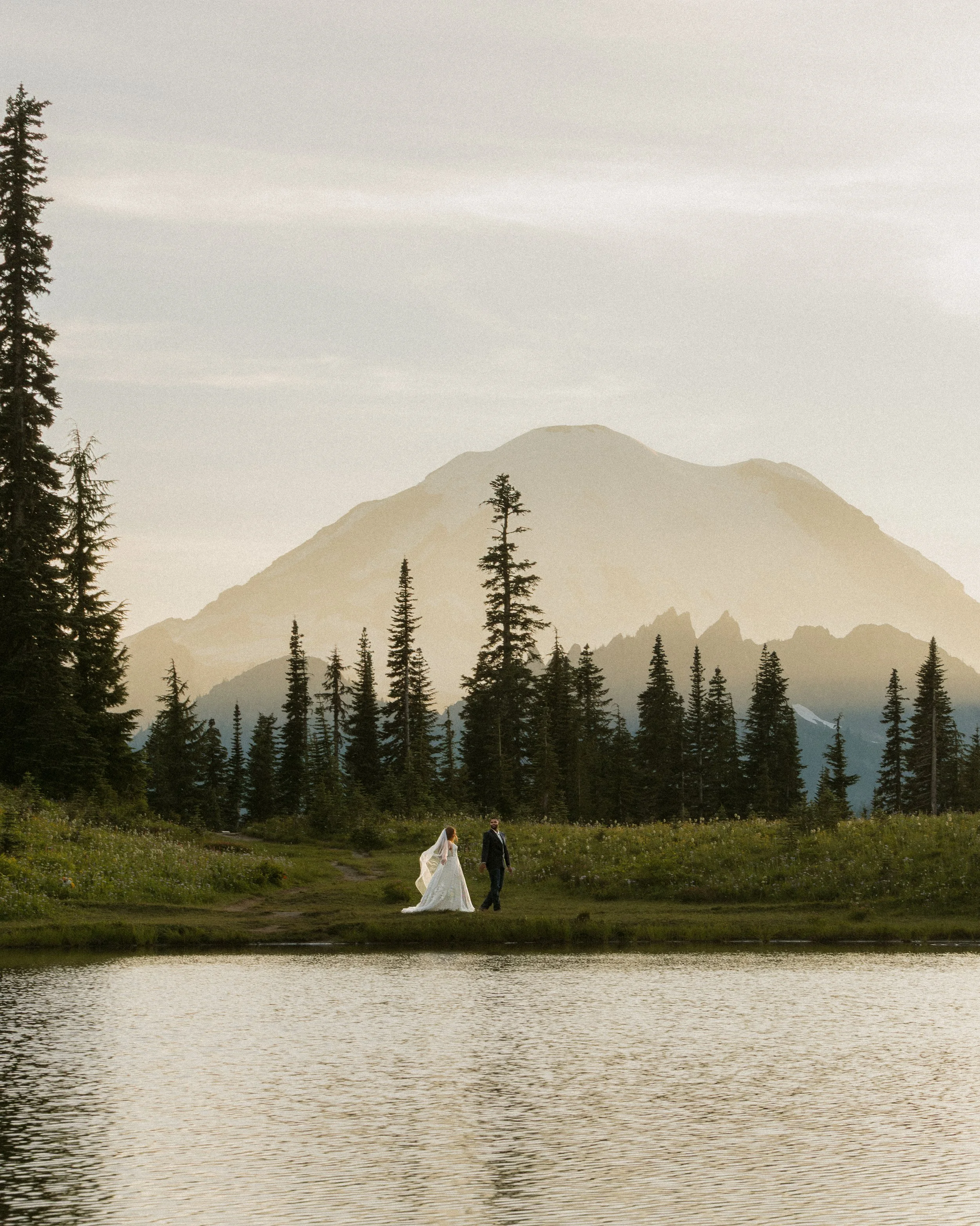 A bride and groom walking near a lake with a mountain and tall pine trees in the background during sunset or sunrise.