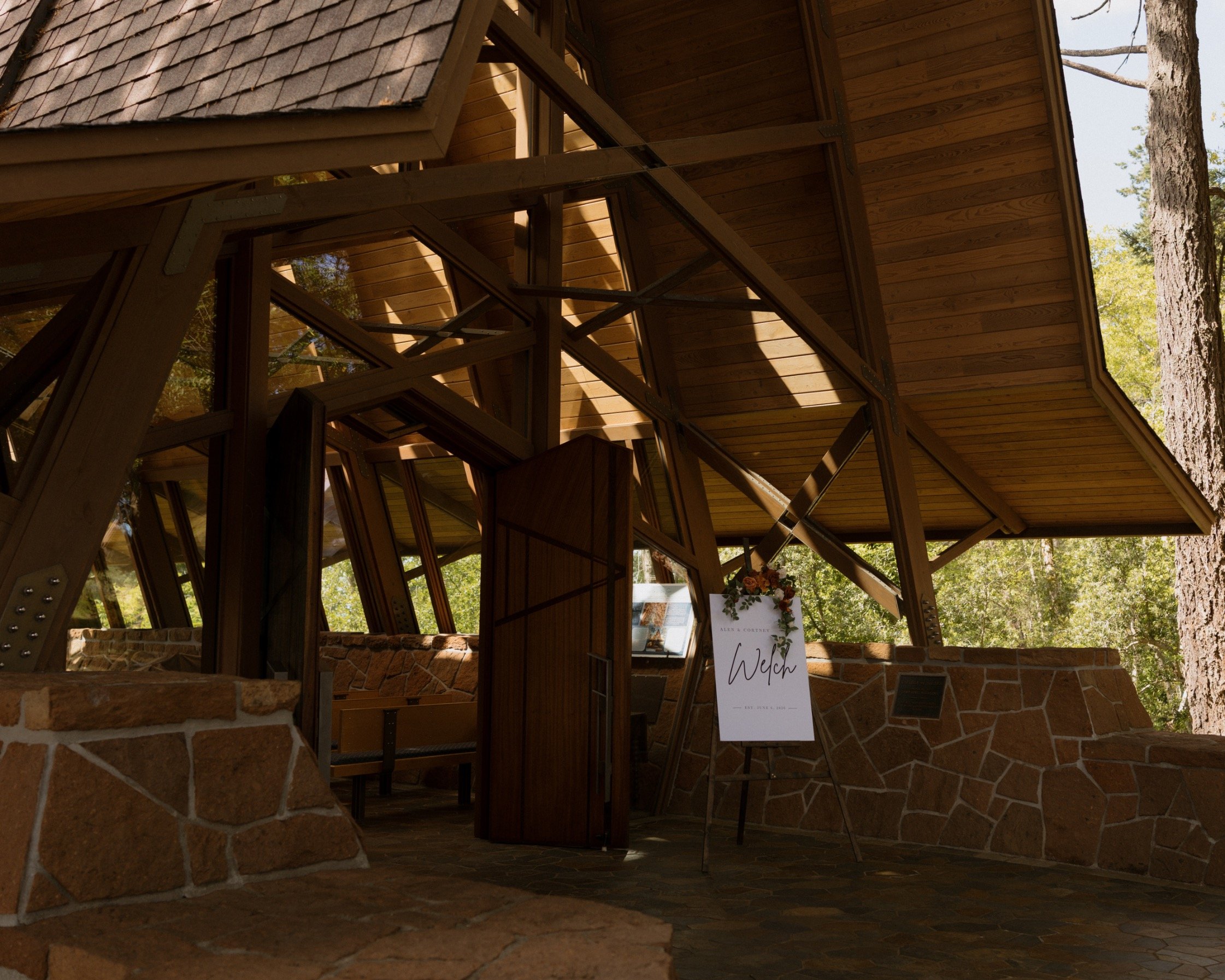 Close-up of a wooden structure with glass windows, supported by metal beams, in an outdoor environment with trees in the background. A sign with the word 'Welch' is partially visible in the lower right corner.