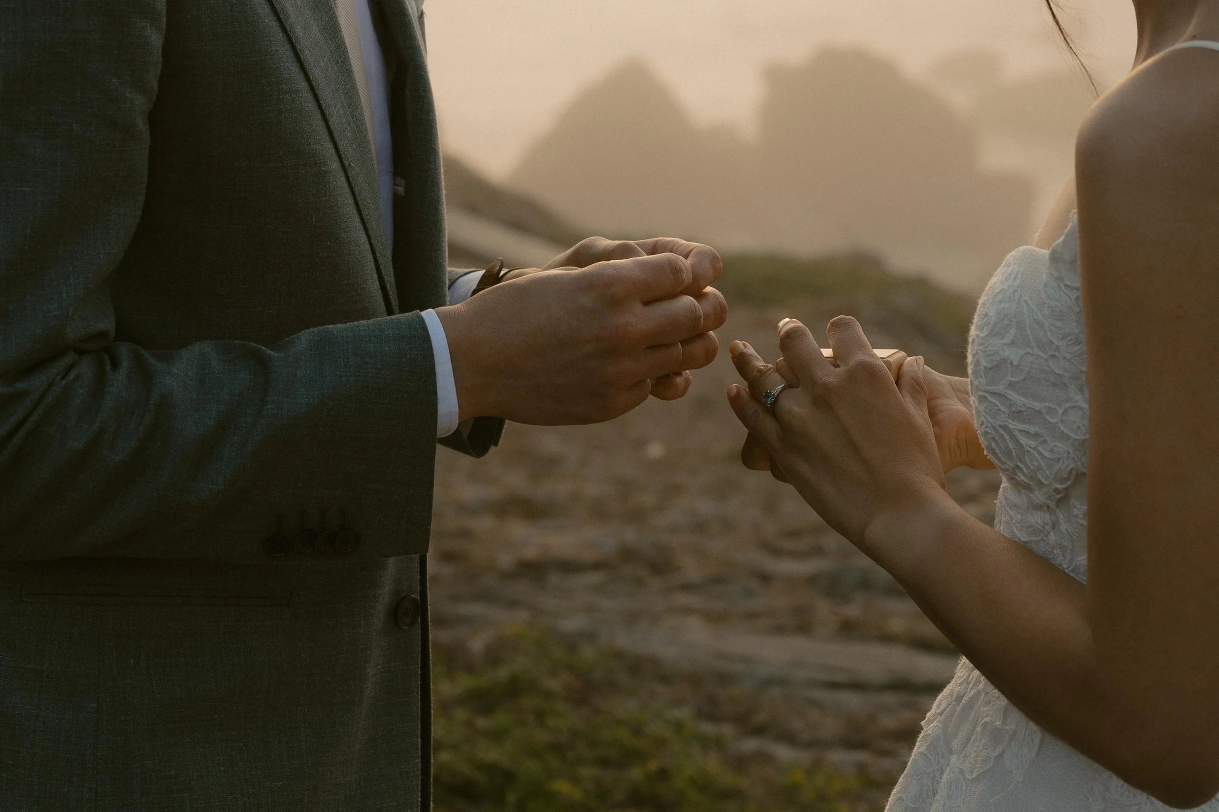 A couple exchanging rings during a wedding ceremony outdoors at sunset, with the focus on their hands and rings.