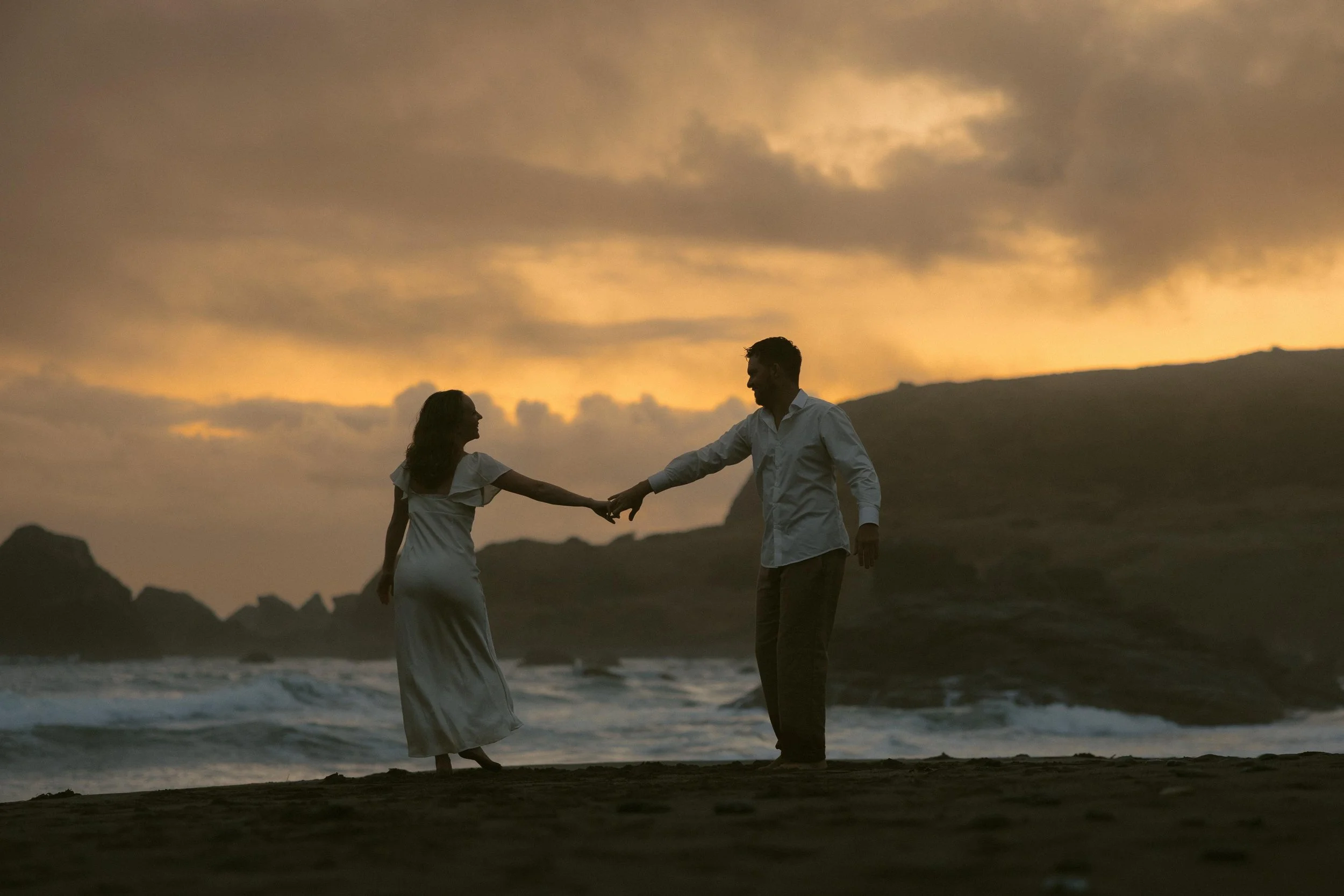 A couple holding hands on the beach at sunset, with mountains in the background and a cloudy sky overhead.