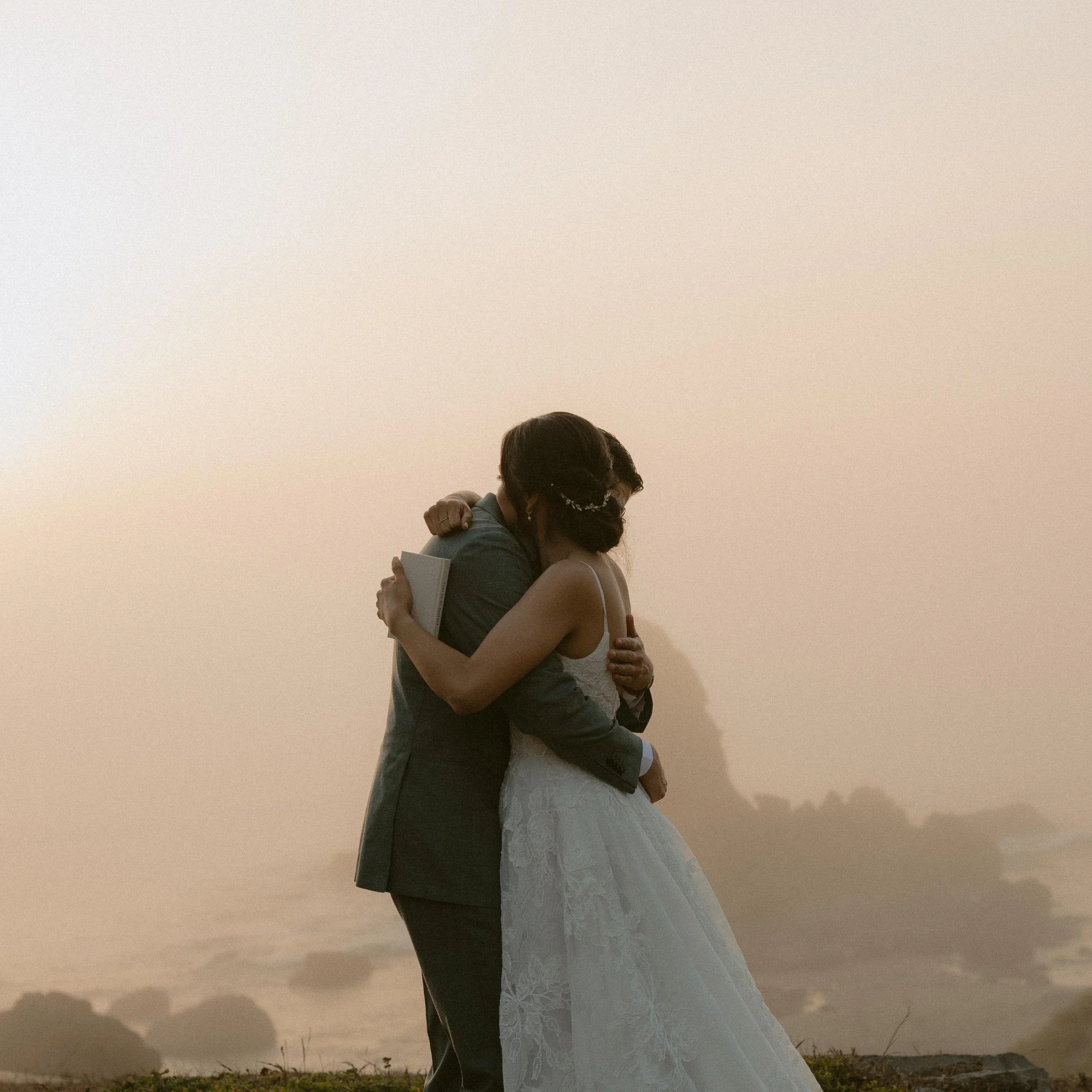 A couple embracing outdoors at dusk, with the woman in a wedding dress and the man in a suit, near a large beige wall.