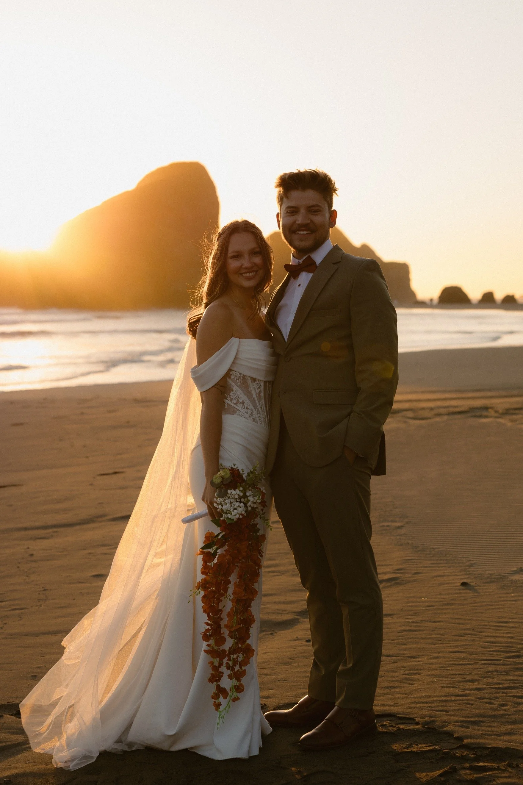 A couple in wedding attire standing on a beach during sunset, with large rock formations in the background. The woman is wearing a white wedding gown and holding a bouquet, and the man is dressed in a gray suit with a bow tie. Both are smiling and lo