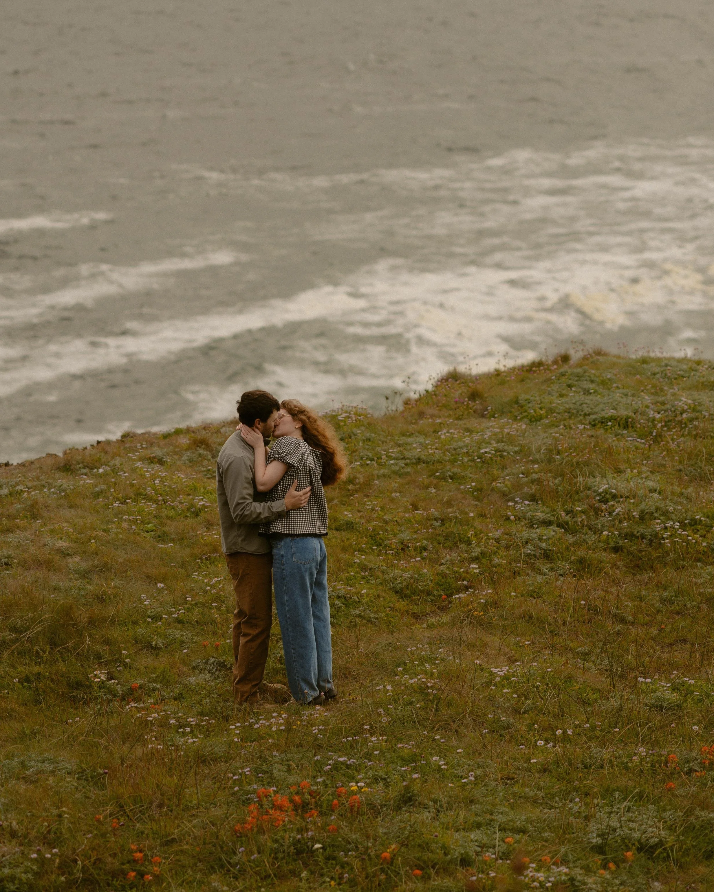 A couple sharing a kiss on a grassy hill with wildflowers, ocean waves, and a cloudy sky in the background.