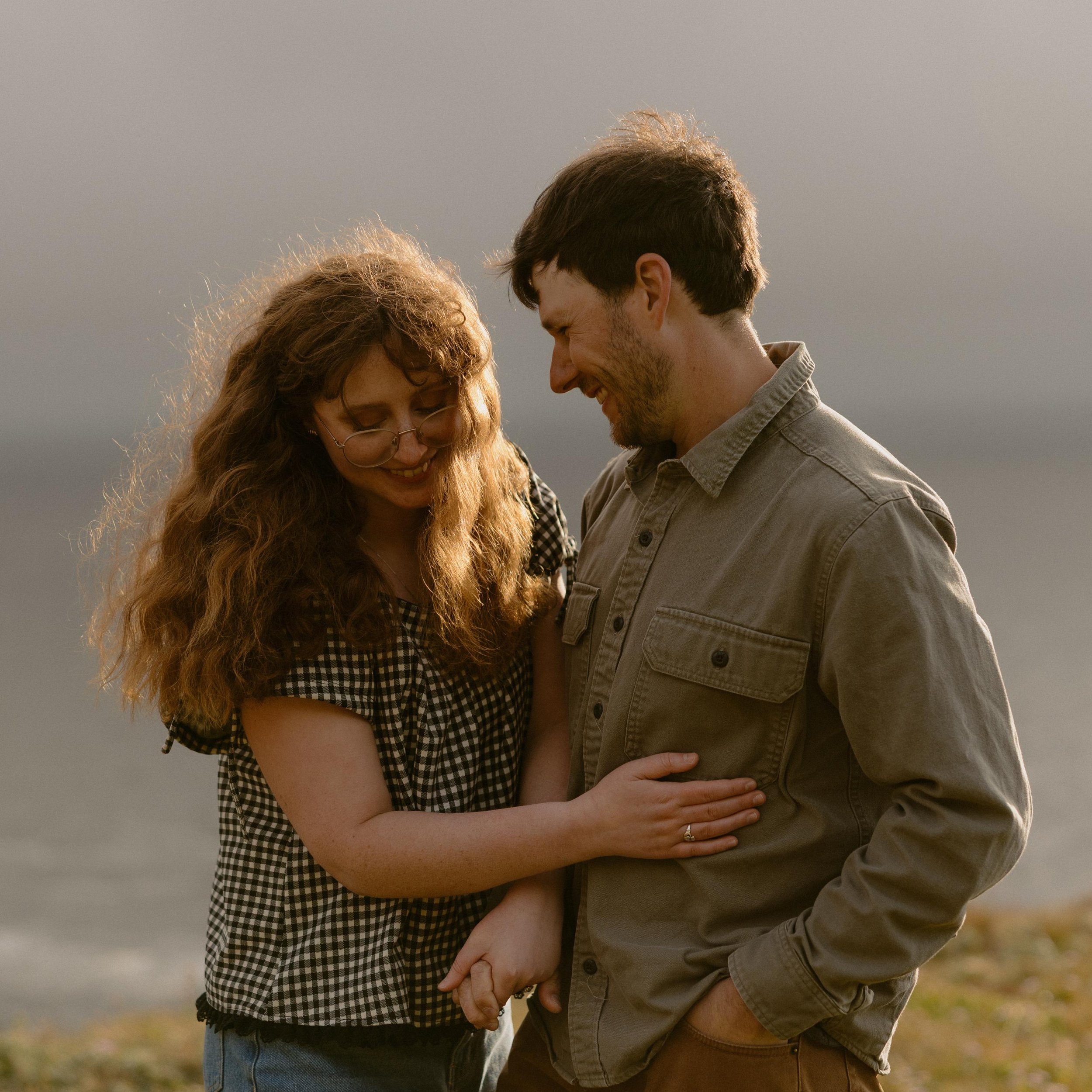 A couple standing close together outdoors, smiling and touching foreheads, holding hands with fingers intertwined, sunset lighting, blurry background.
